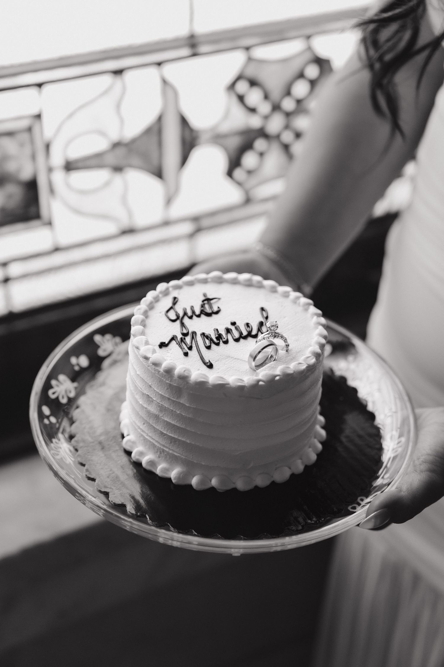 Black and white photo of a small round wedding cake reading Just Married on a glass plate held by a bride.