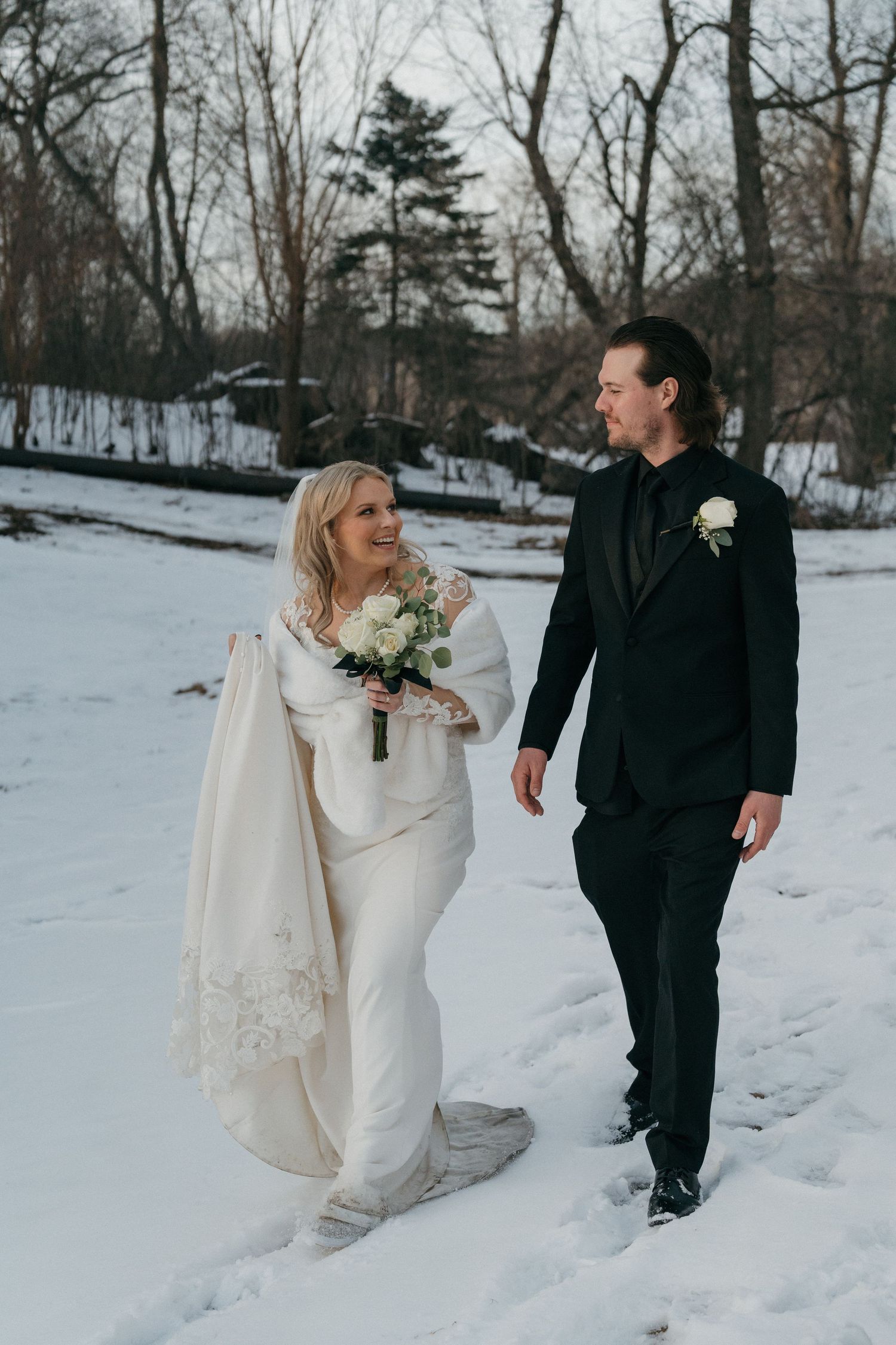 Bride in white gown with fur wrap and groom in black tuxedo walking through a snowy winter landscape in Minnesota.
