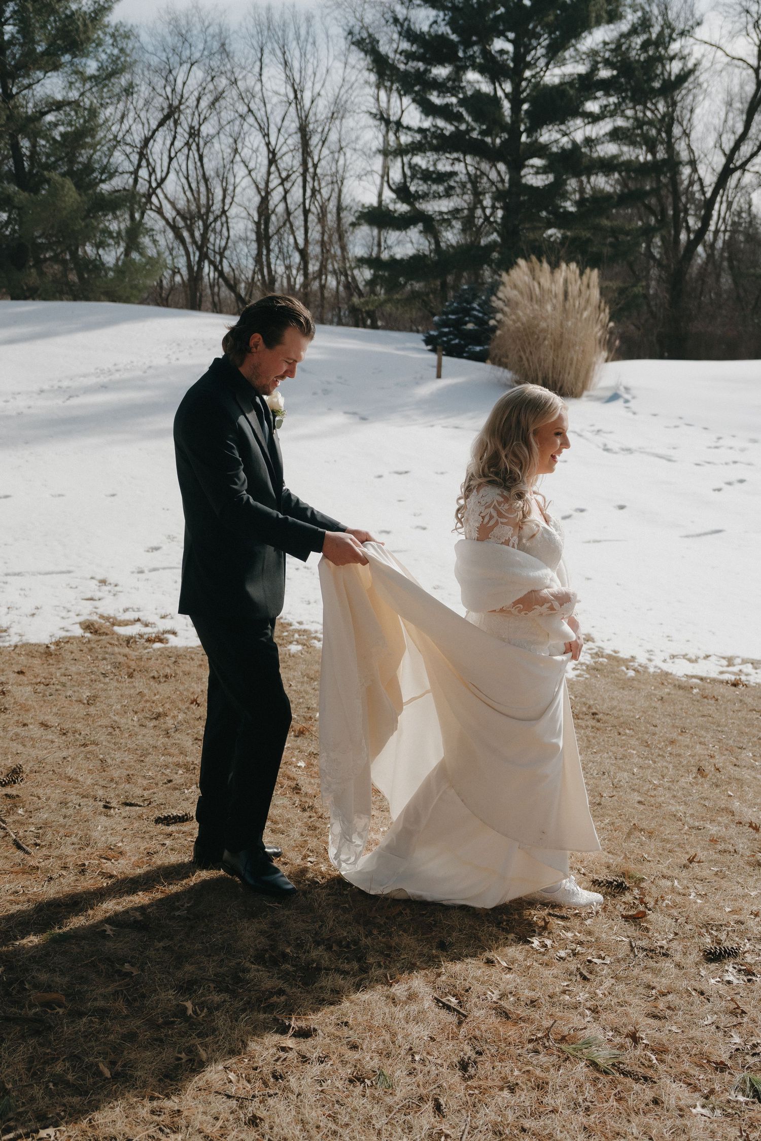 Groom helps arrange bride's flowing wedding gown train in snowy winter outdoor landscape in Minnesota.