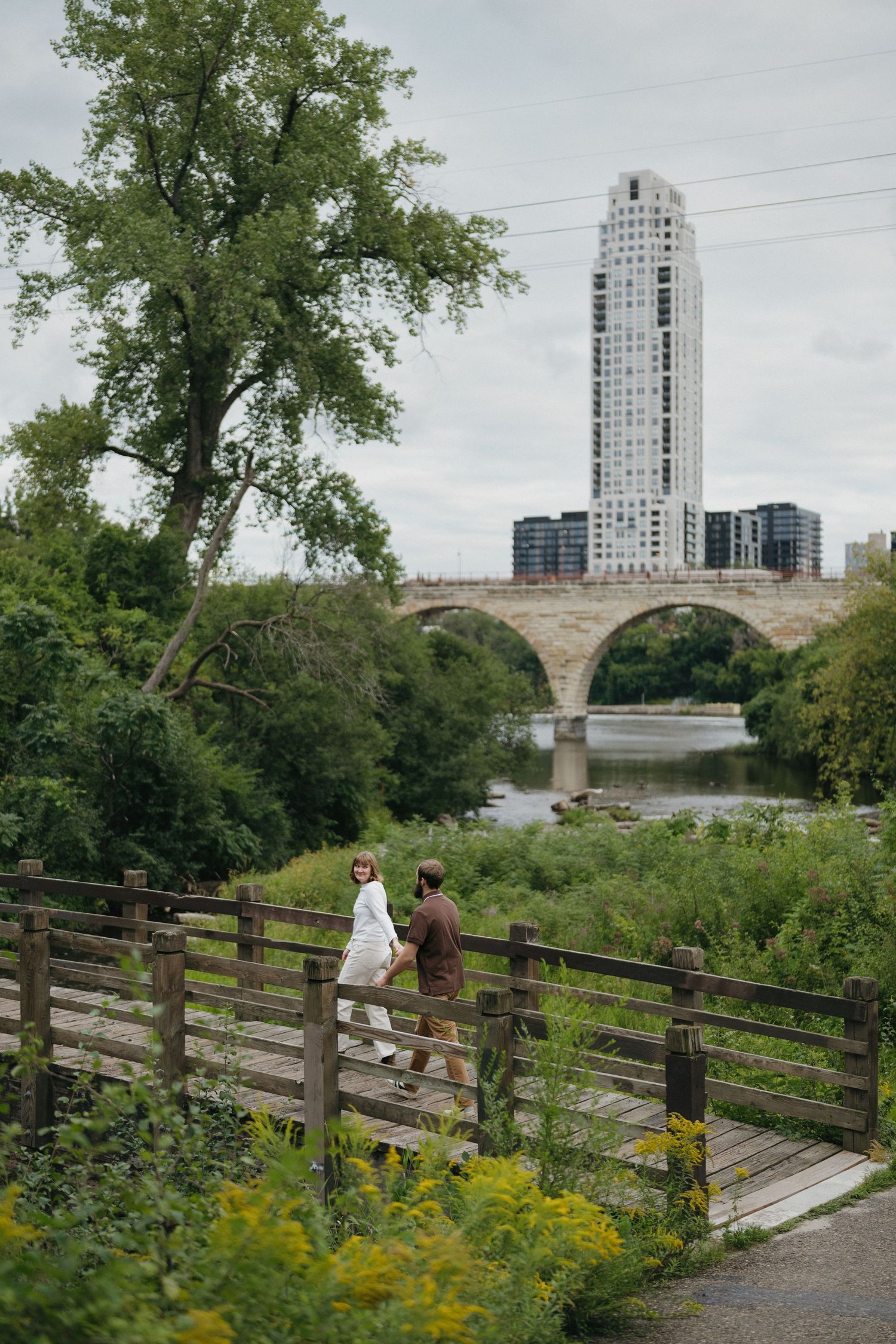 Couple walks on wooden bridge path with stone arch bridge and city skyline behind in Minneapolis, Minnesota.