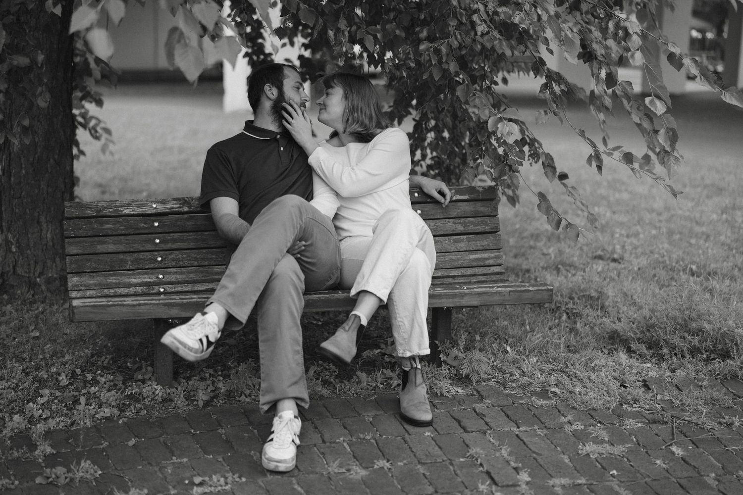 Black and white photo of a couple sharing a kiss on a park bench under a tree near Saint Anthony Main in Minneapolis.