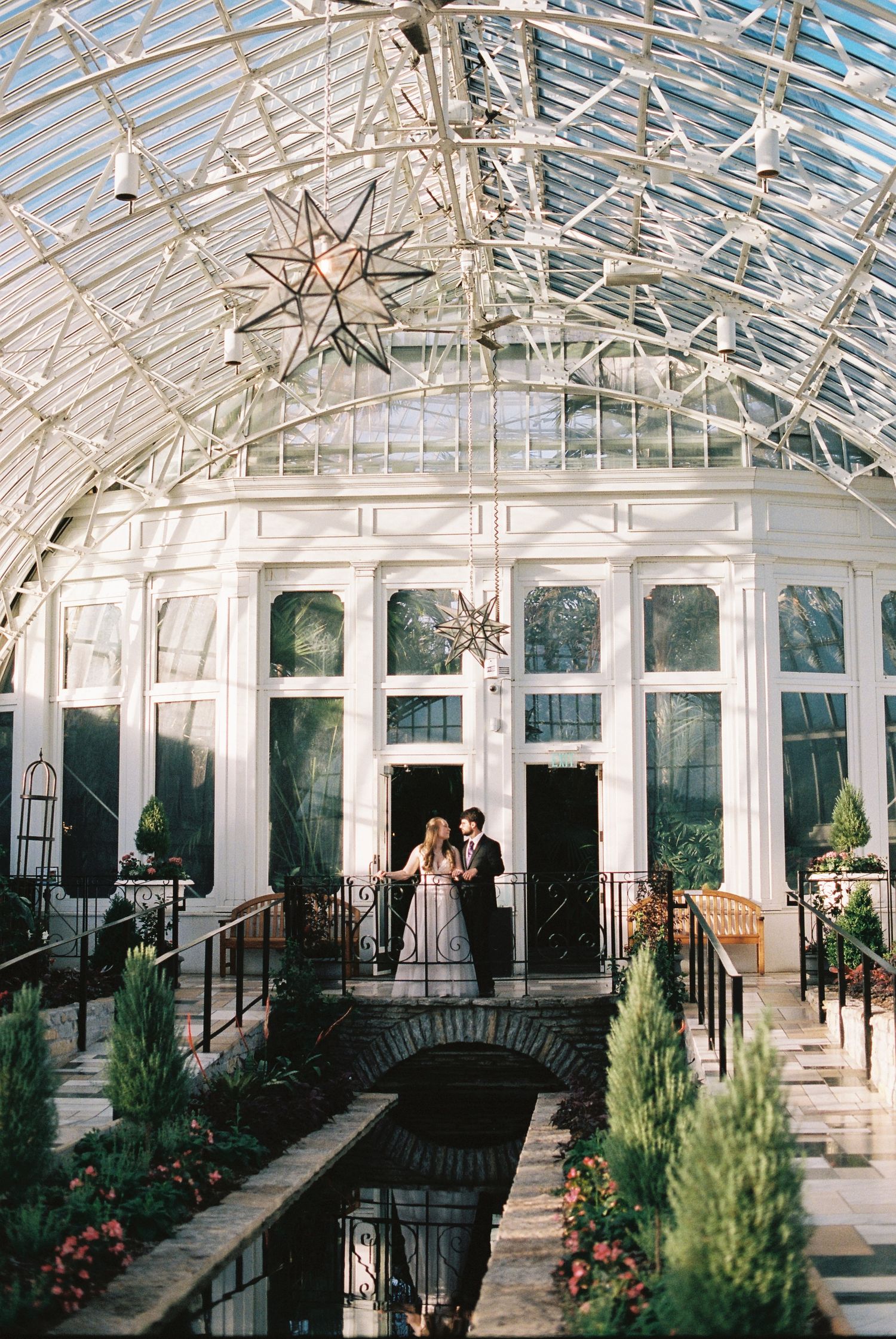 Couple embraces on a bridge inside a stunning glass conservatory greenhouse wedding venue with star chandeliers.