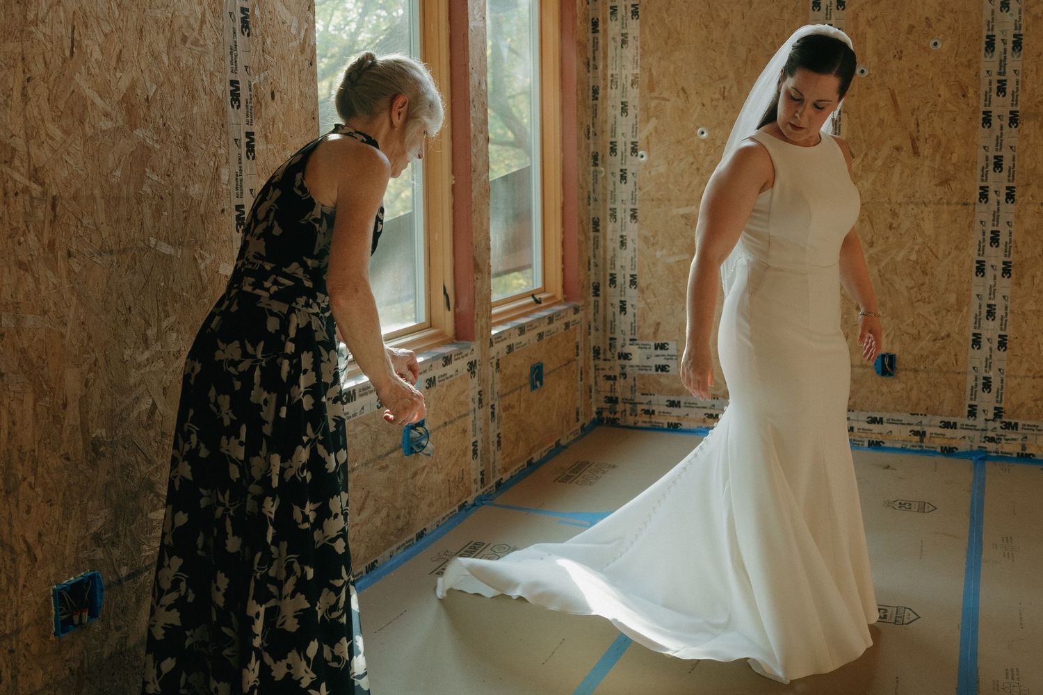 Mother of bride in floral dress adjusting bride's elegant white gown train in rustic unfinished room in Southern Minnesota.