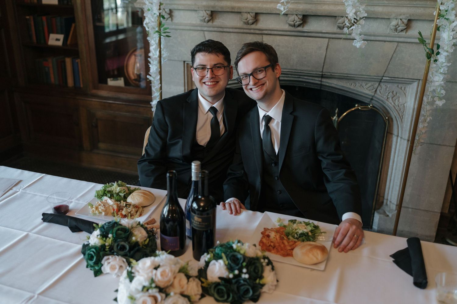 Two grooms in black suits smile together at their wedding reception table with wine and flowers in Rochester, Minnesota.