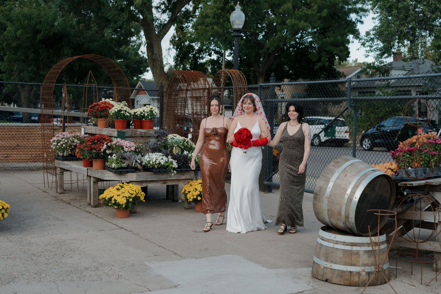 Bride with red bouquet poses with two bridesmaids outside rustic outdoor greenhouse at Mother Earth Gardens in Longfellow, Minnesota.