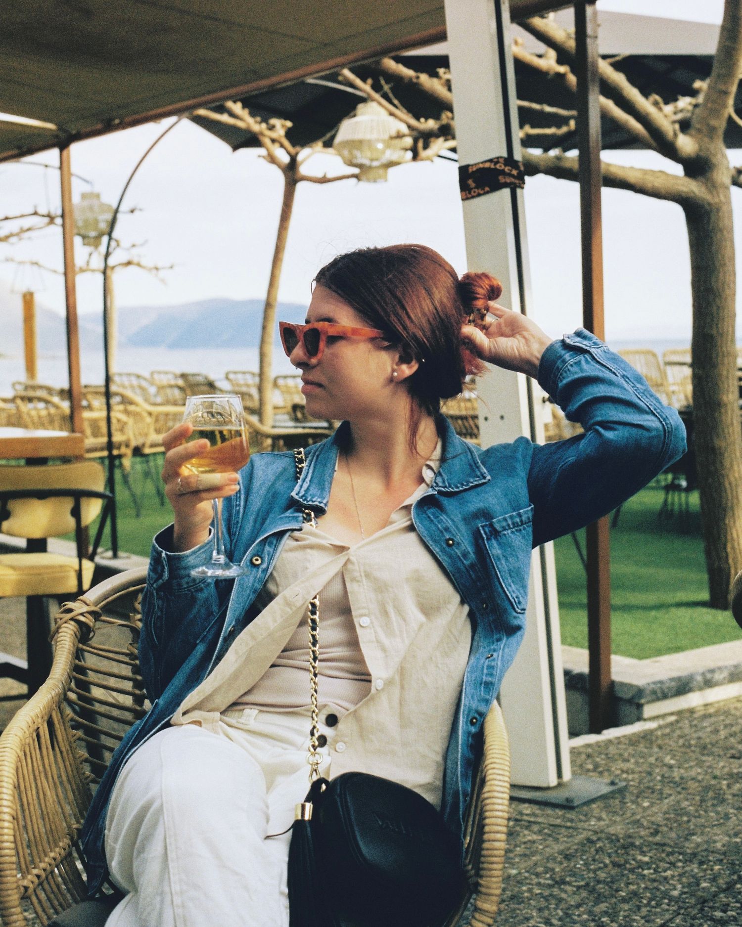Woman in denim jacket and red sunglasses enjoying a drink at an outdoor cafe with scenic water view in Greece.