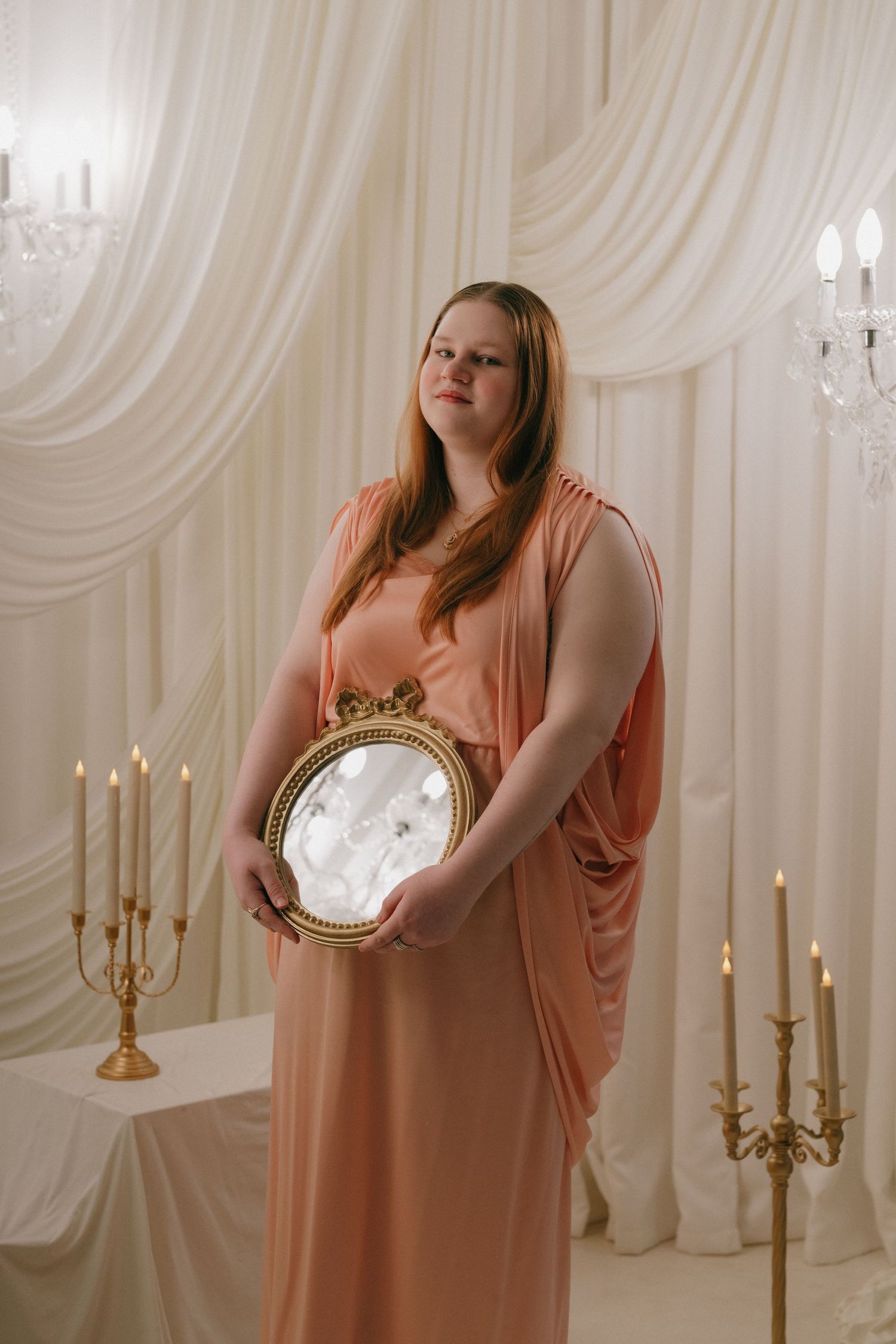 A plus-size woman in a peach gown holds a decorative gold mirror in an elegant candlelit setting in a studio in Saint Paul, Minnesota.