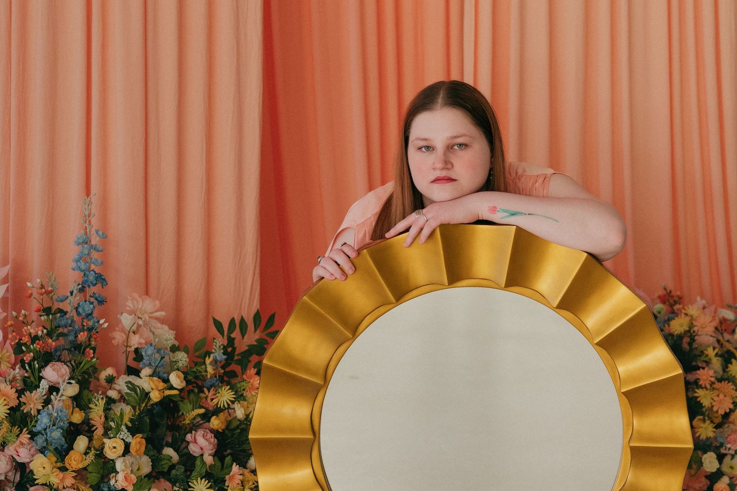 Woman leans on large gold sunburst mirror surrounded by colorful flowers against peach curtain backdrop in a studio setting in St. Paul, MN.