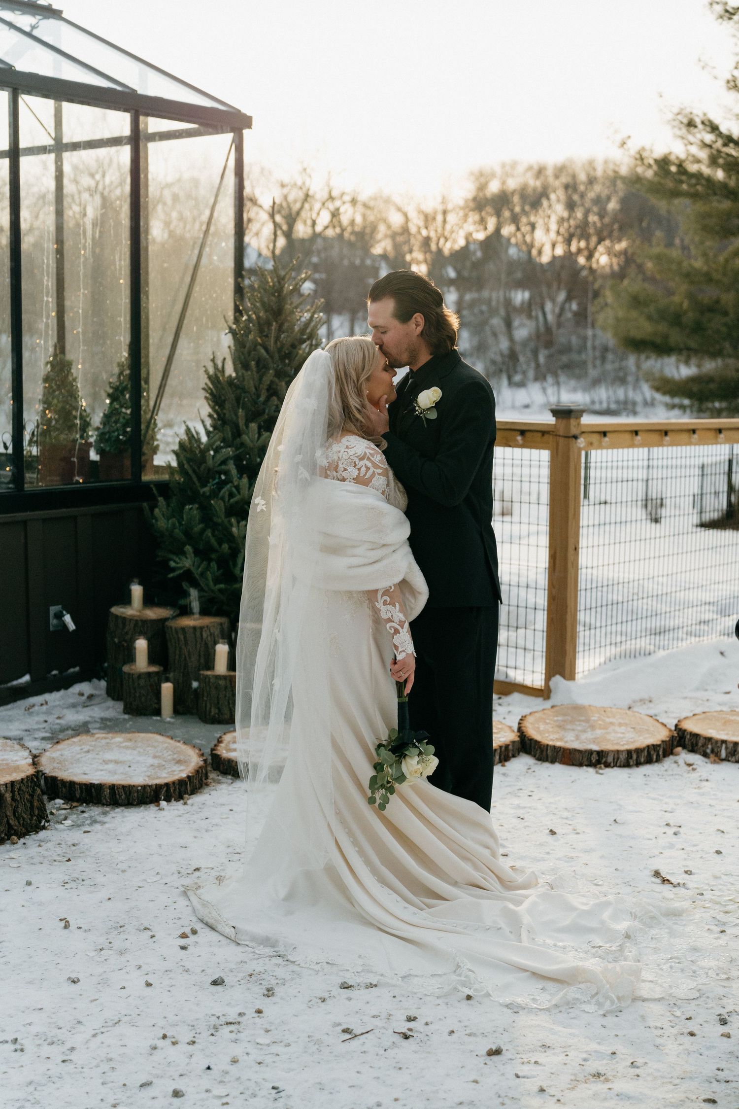 Bride and groom share a kiss outdoors in a snowy winter wedding setting with candles and wood stumps in Minnesota.