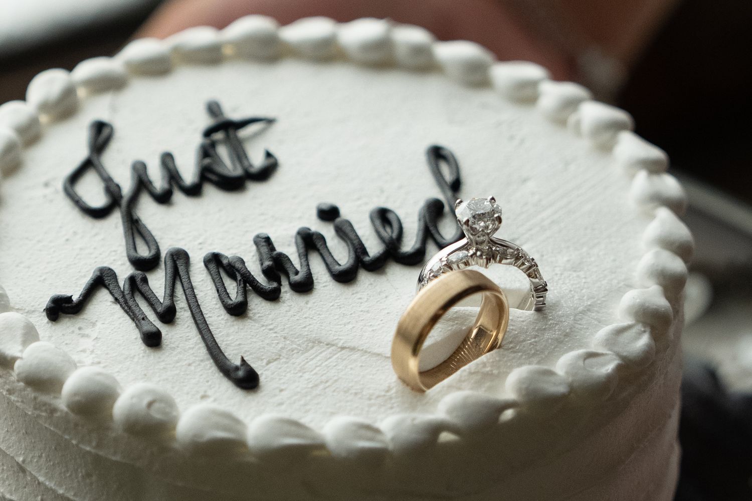 Close-up of a Just Married wedding cake topped with a diamond engagement ring and gold wedding band.