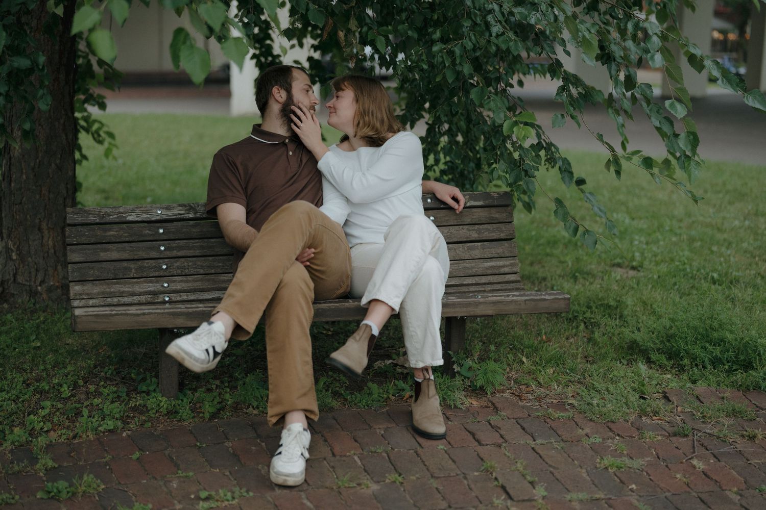 Couple sharing a kiss on a wooden park bench under green tree branches near St. Anthony Main in Minneapolis.