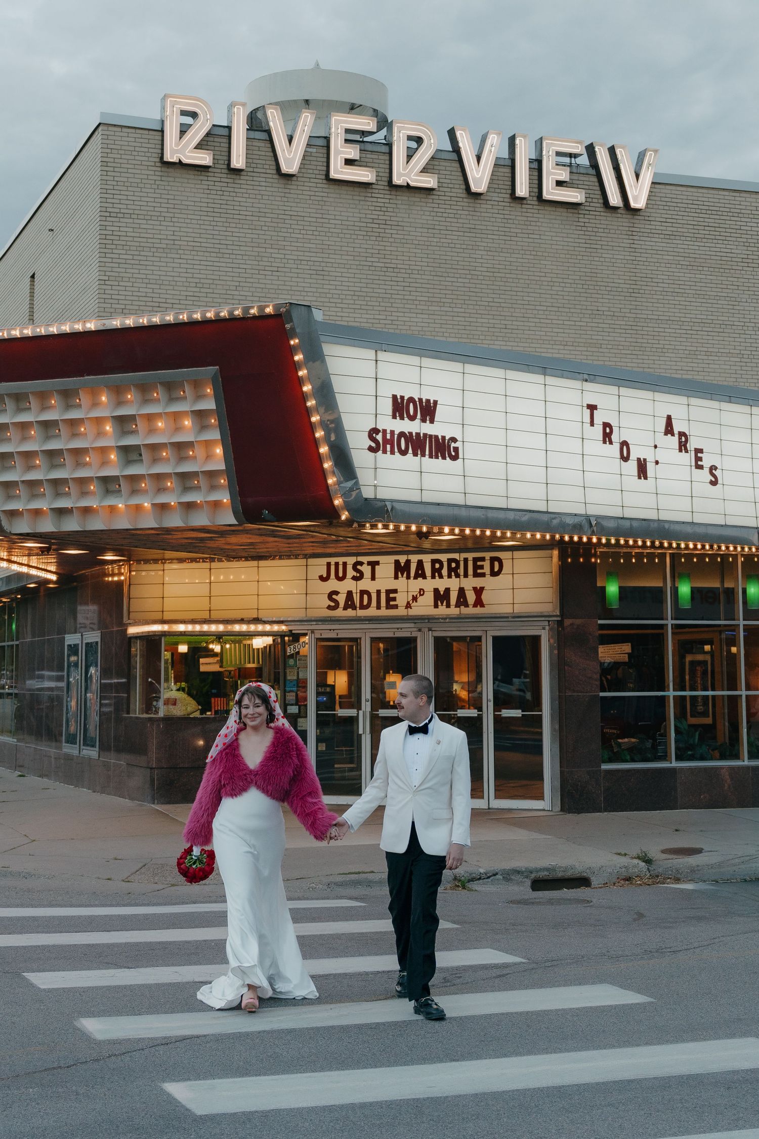 Just married couple walks hand-in-hand across crosswalk in front of Riverview Theater marquee in Minneapolis, Minnesota.