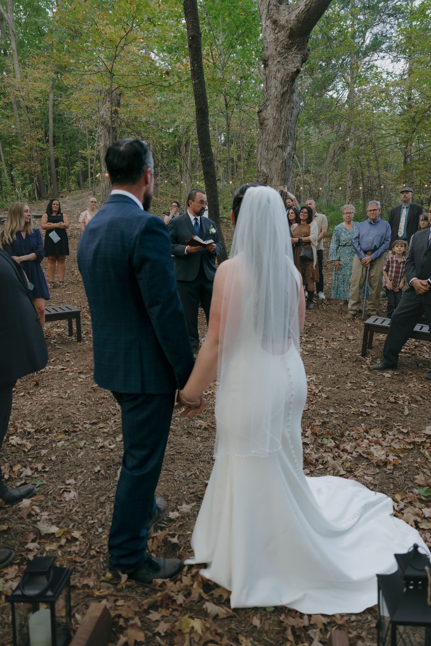 Bride and groom hold hands during an outdoor woodland wedding ceremony surrounded by guests among autumn leaves.
