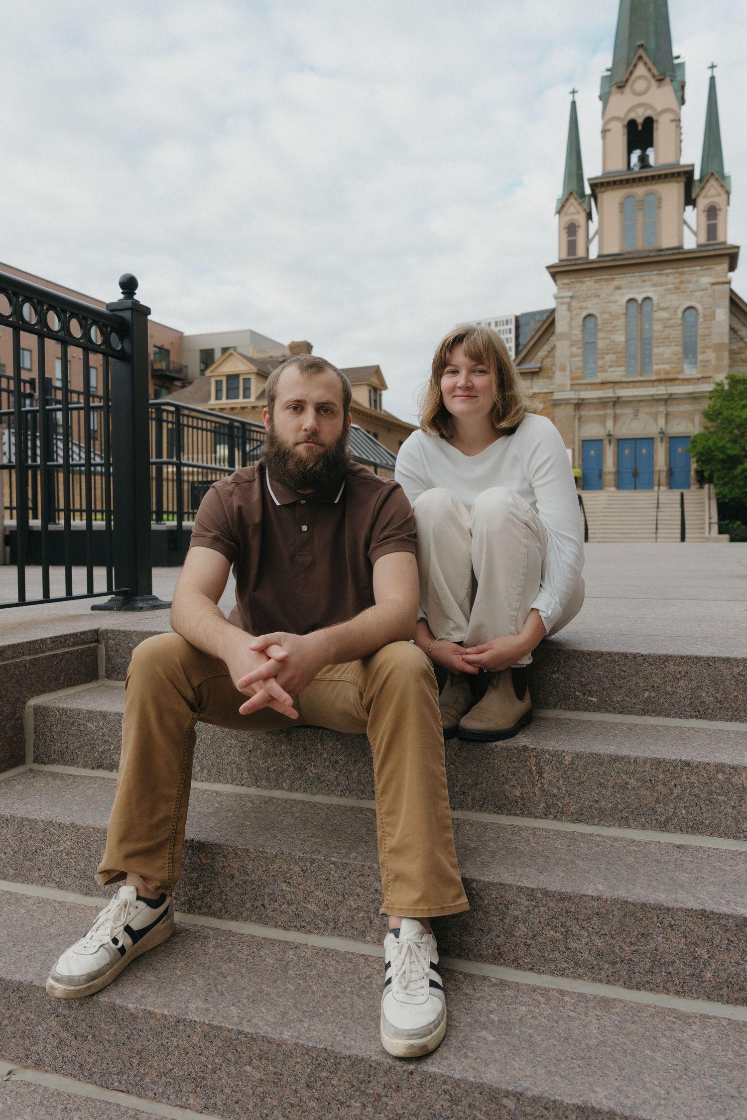 Couple in casual brown and white outfits sitting on stone steps with church in background near St. Anthony Main in Minneapolis.