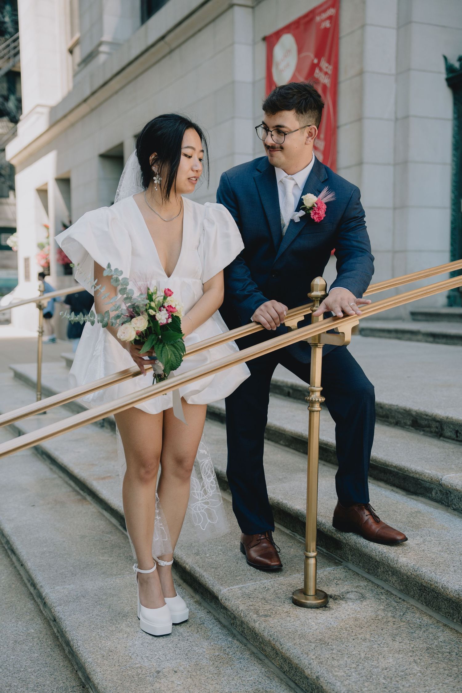 Asian bride in short white dress holds bouquet beside groom in navy suit on urban building steps in Chicago, Illinois.