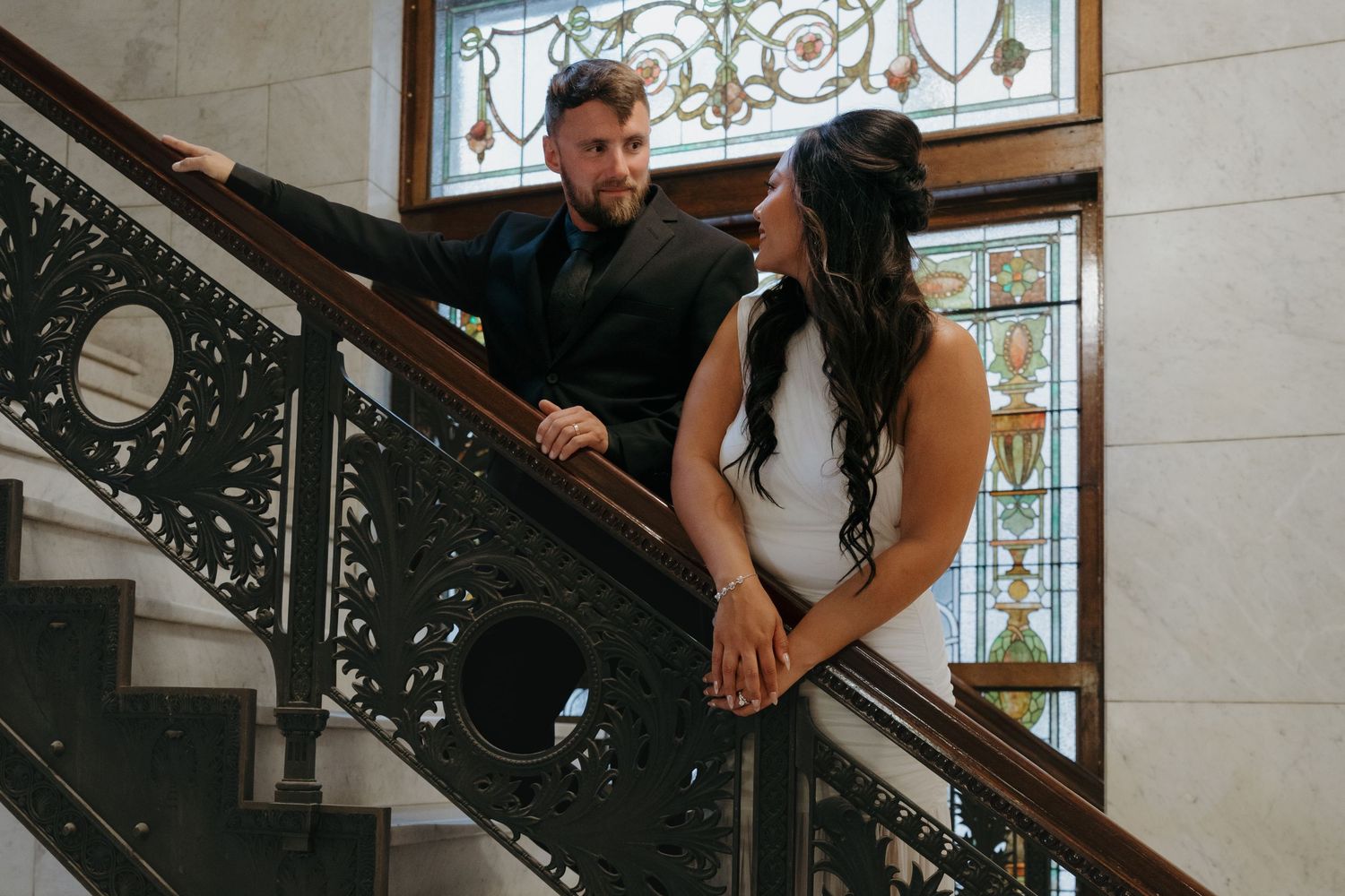 Couple shares romantic moment on decorative iron staircase with stained glass window backdrop at Minneapolis City Hall.