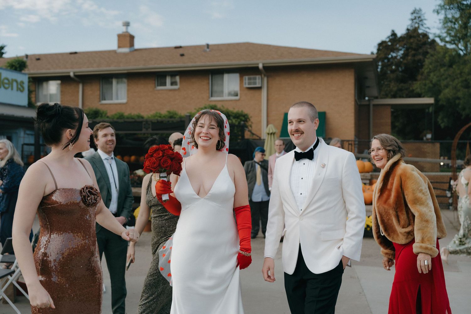 Smiling bride in white gown and groom in white tuxedo walking with wedding guests at outdoor fall celebration.