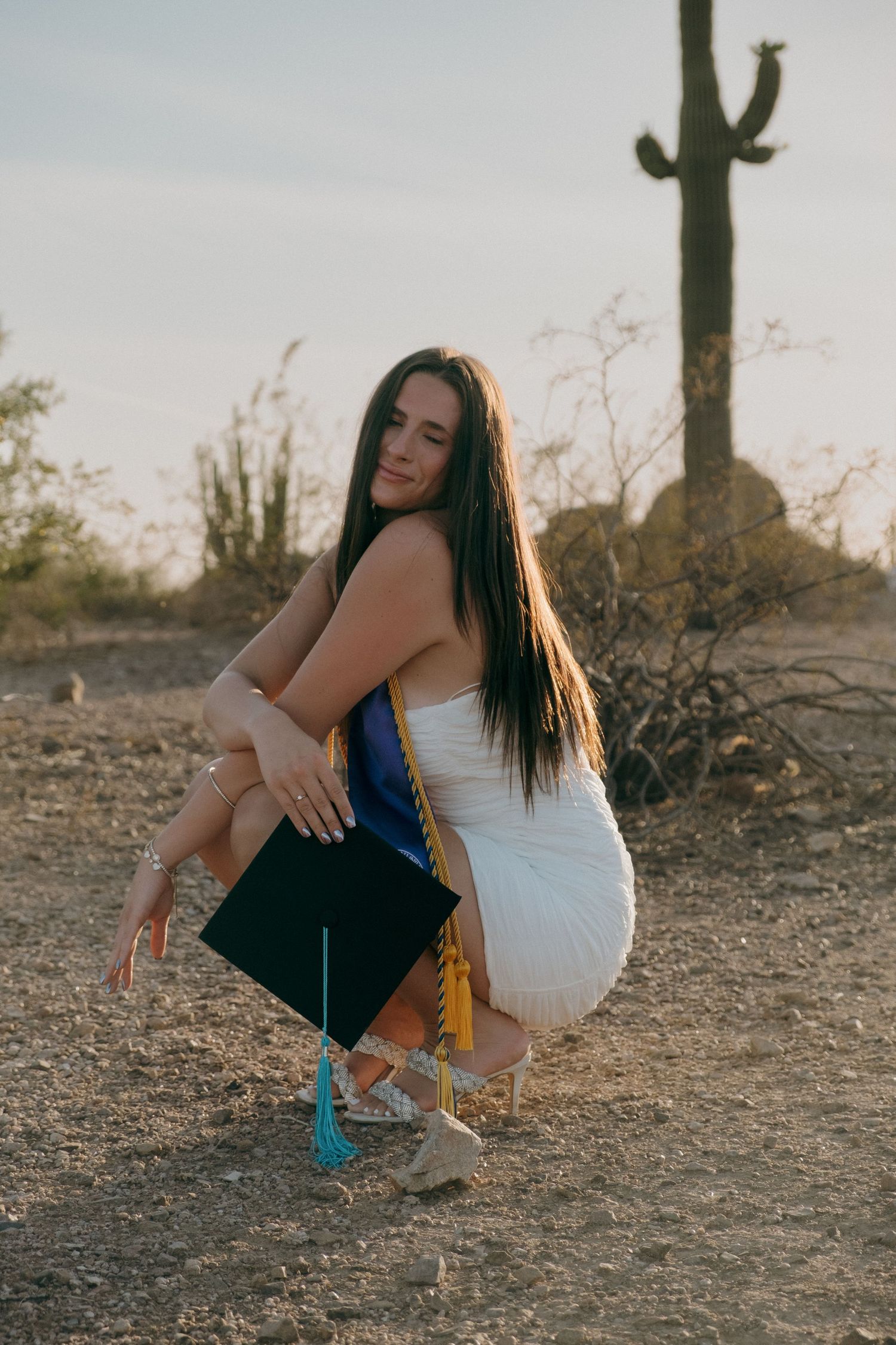 Graduate in white dress holding cap and sash crouching in Arizona desert setting near Phoenix, Arizona.