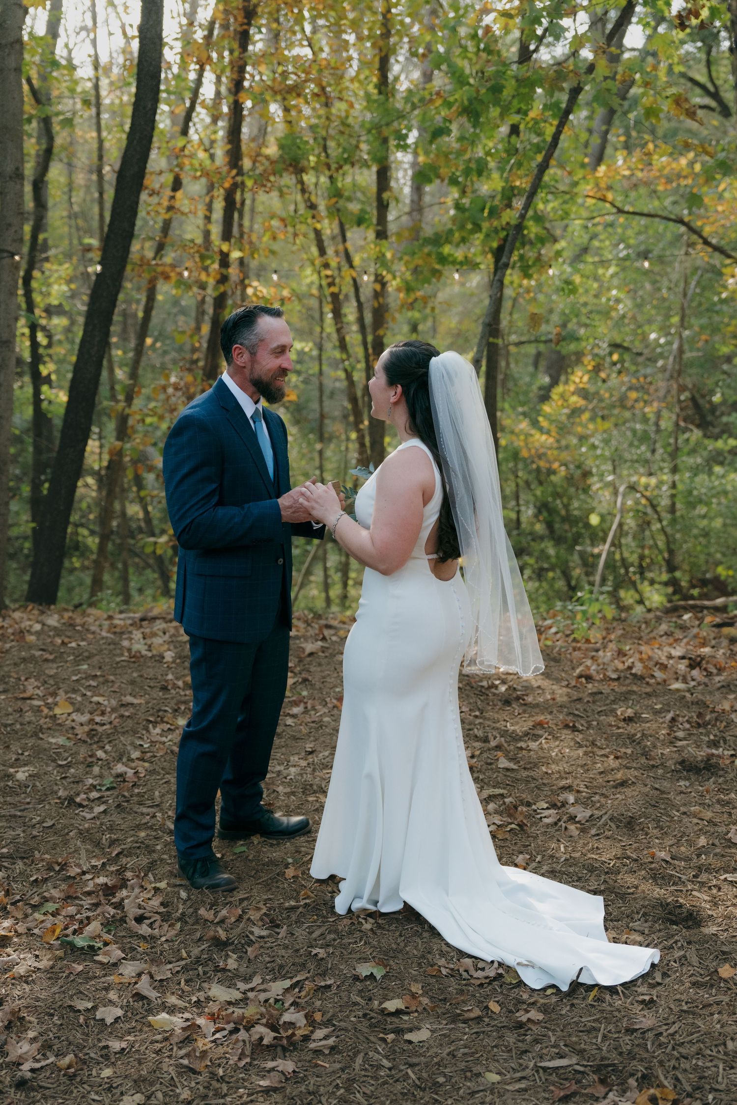 Bride in white gown with veil holds hands with groom in navy suit in autumn forest setting in Shakopee, Minnesota.