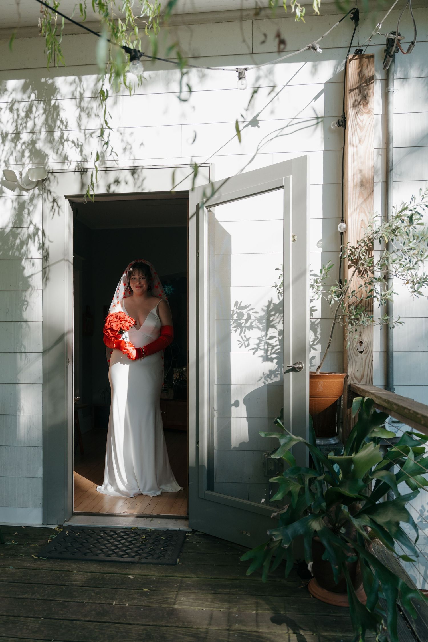 Bride in white gown with red bouquet stands in a sunlit doorway of a charming white wooden home.