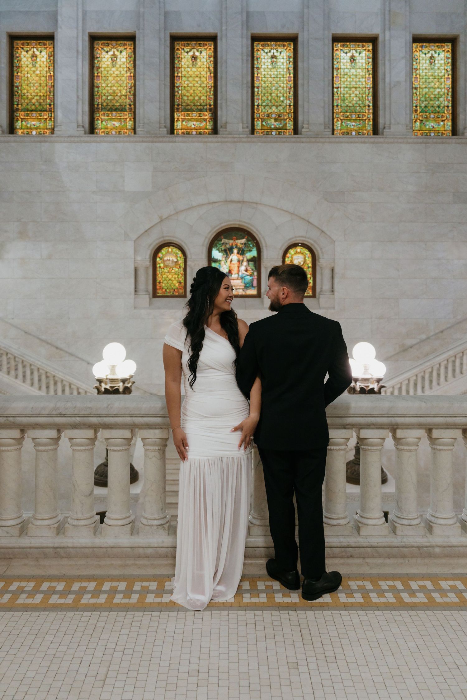 Bride and groom hold hands facing colorful stained glass windows in grand marble hall located in Minneapolis City Hall.