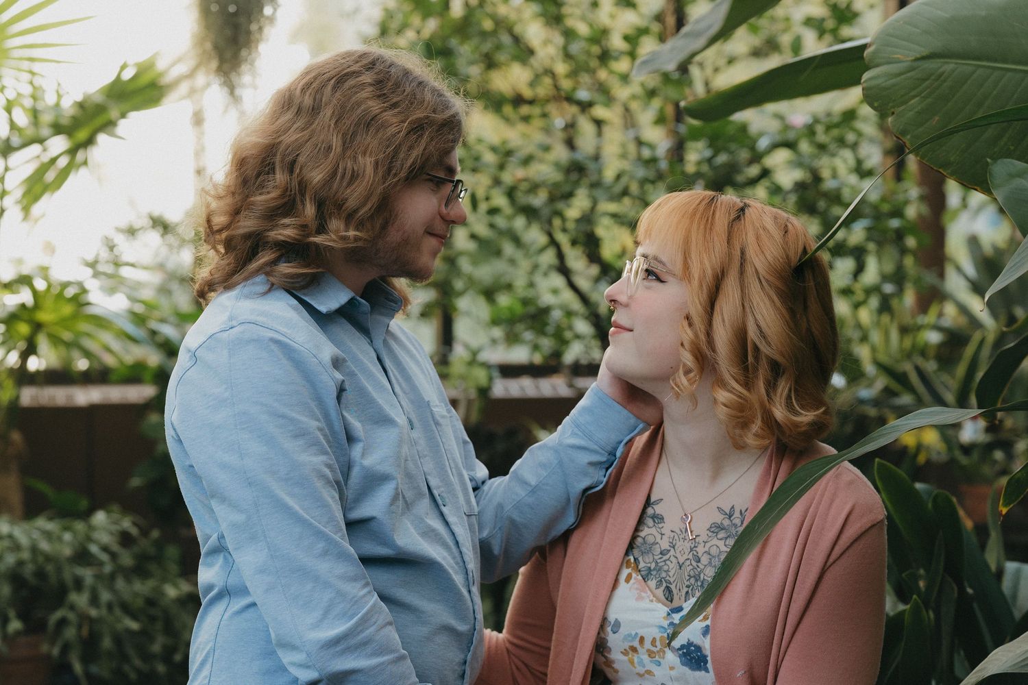 A couple shares a tender moment surrounded by tropical green plants in a sunlit botanical garden at the Minnesota Landscape Arboretum.
