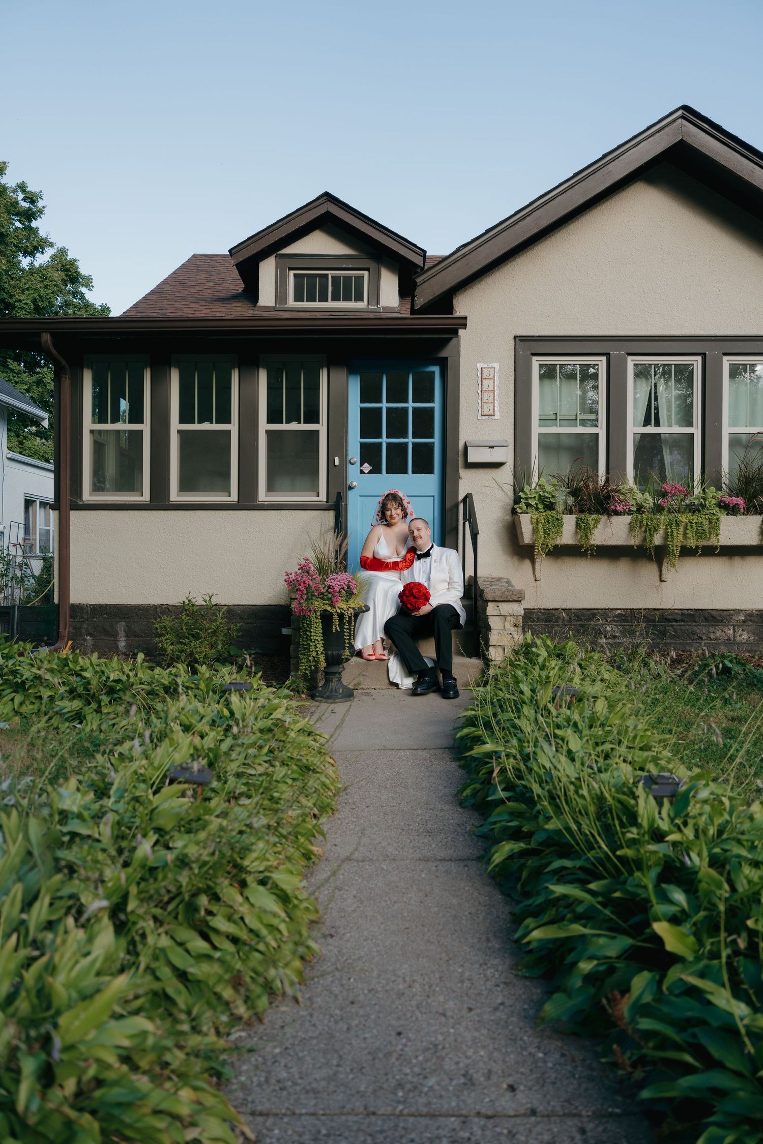 Bride and groom sitting on front porch steps of cozy bungalow home surrounded by lush greenery in Longfellow, Minneapolis, Minnesota.