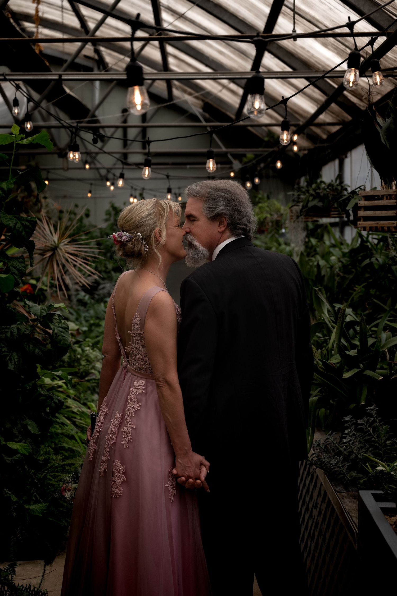 Couple shares romantic kiss in dimly lit greenhouse with Edison bulb string lights overhead.