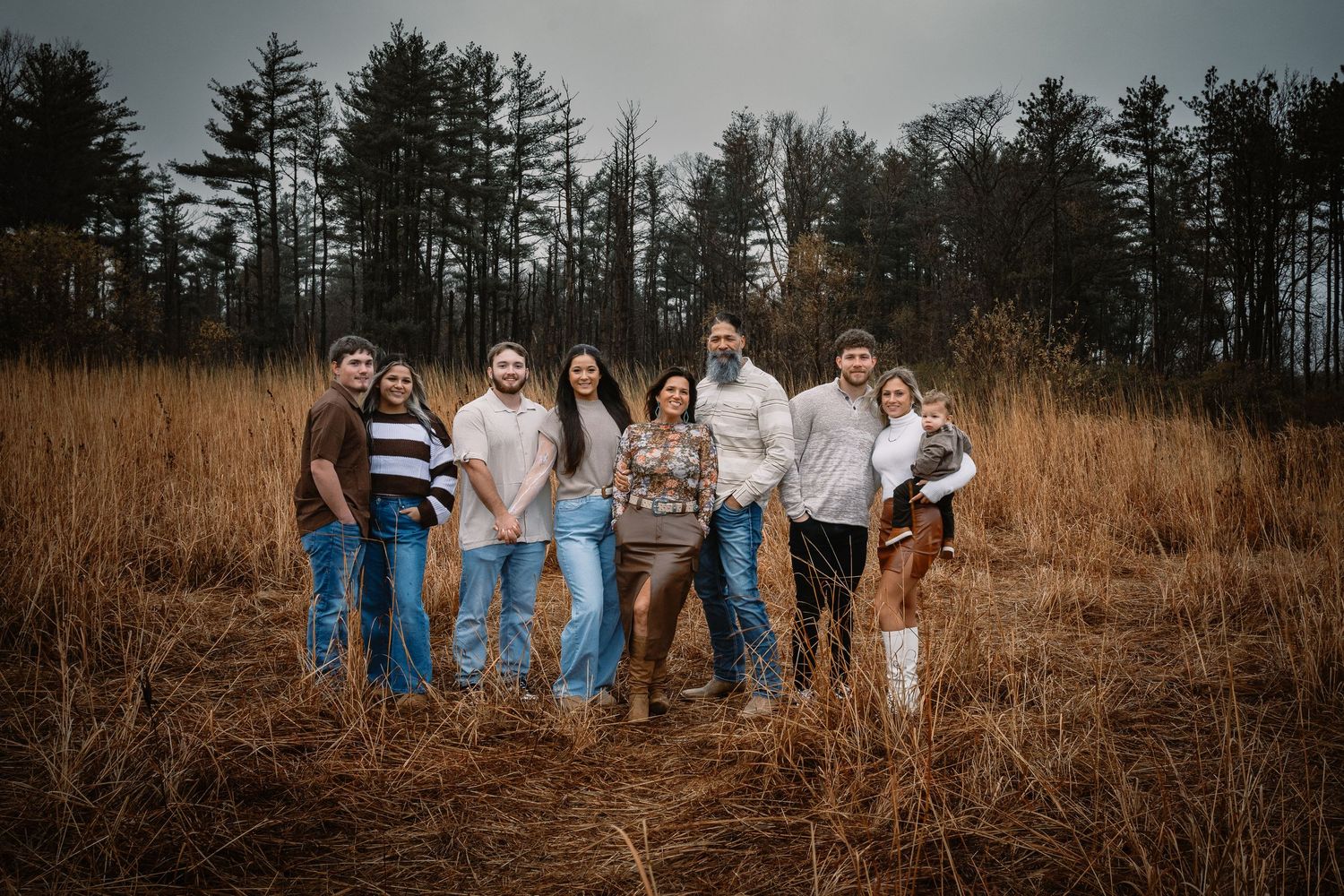 Group of eight people posing together in a dry autumn field with pine tree backdrop.