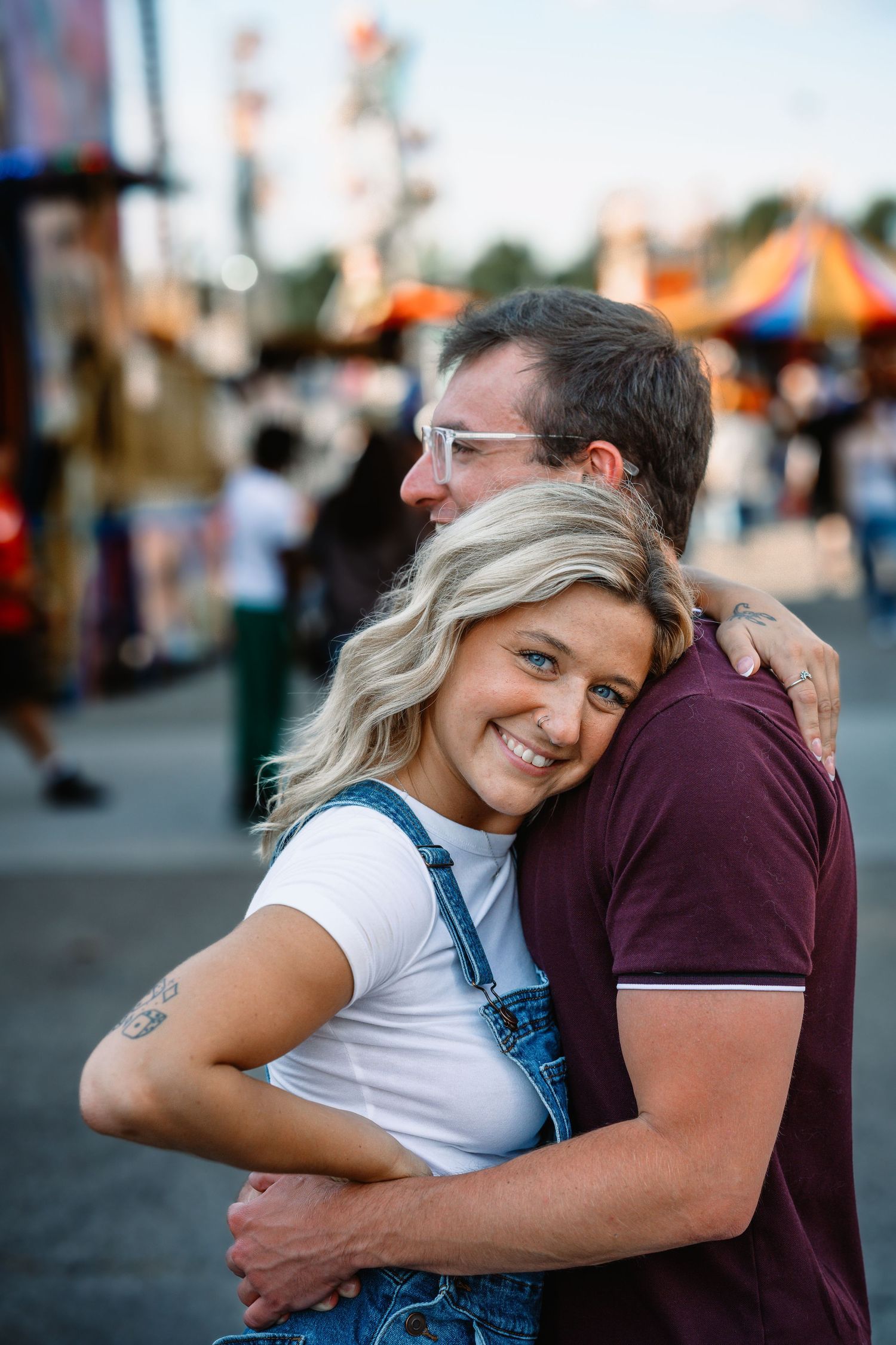Happy blonde woman hugging her partner and smiling at a colorful outdoor carnival.