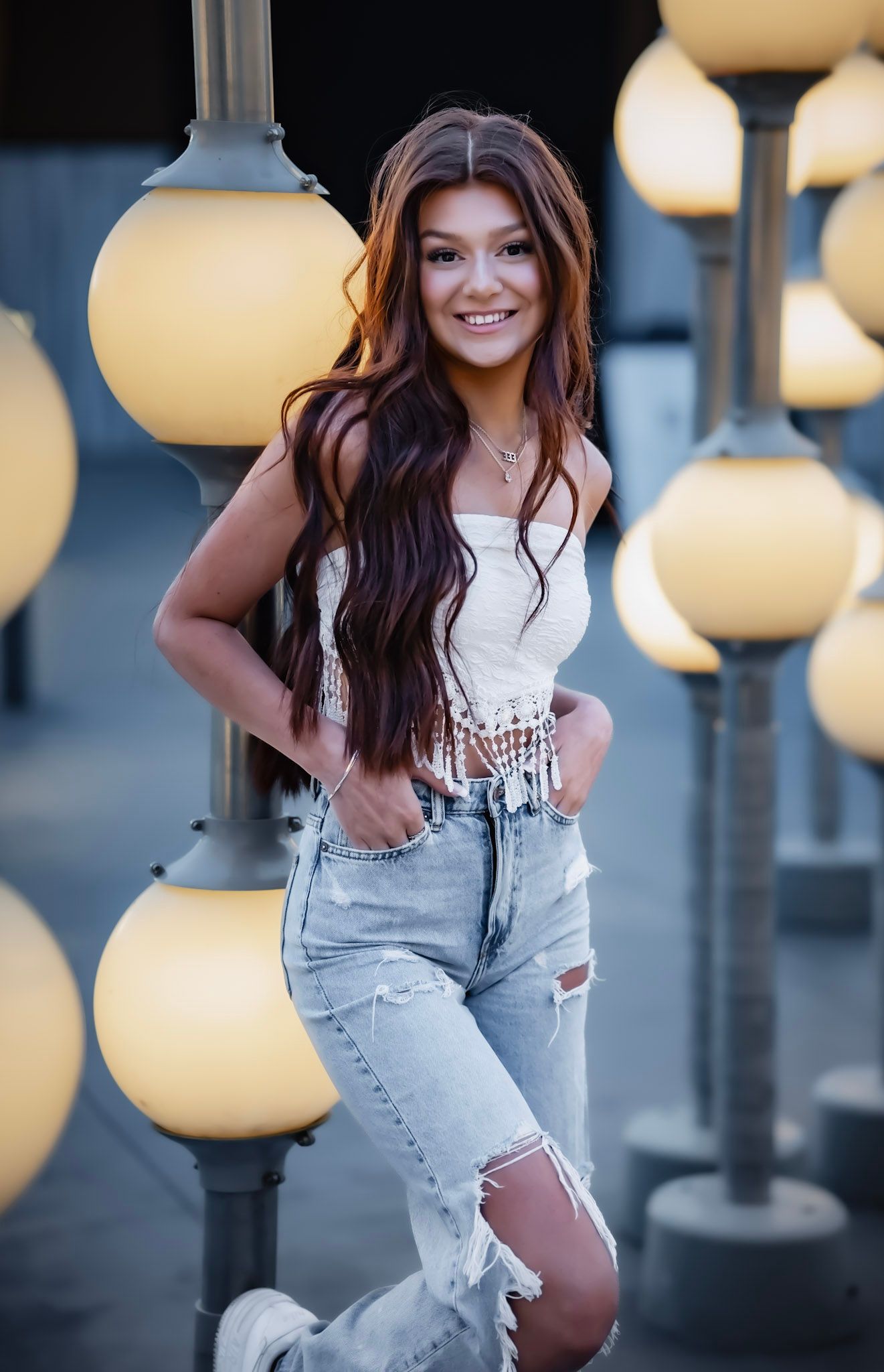 Young woman with long auburn hair smiles in white crop top and ripped jeans, posing among glowing globe lights.