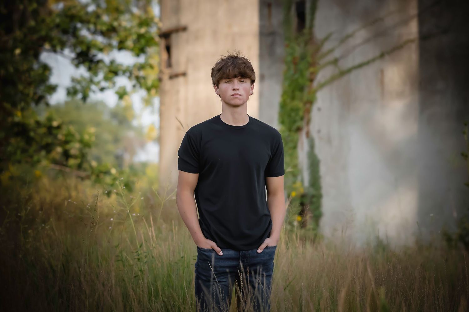 Teen boy in black t-shirt stands in overgrown field near weathered industrial structures.