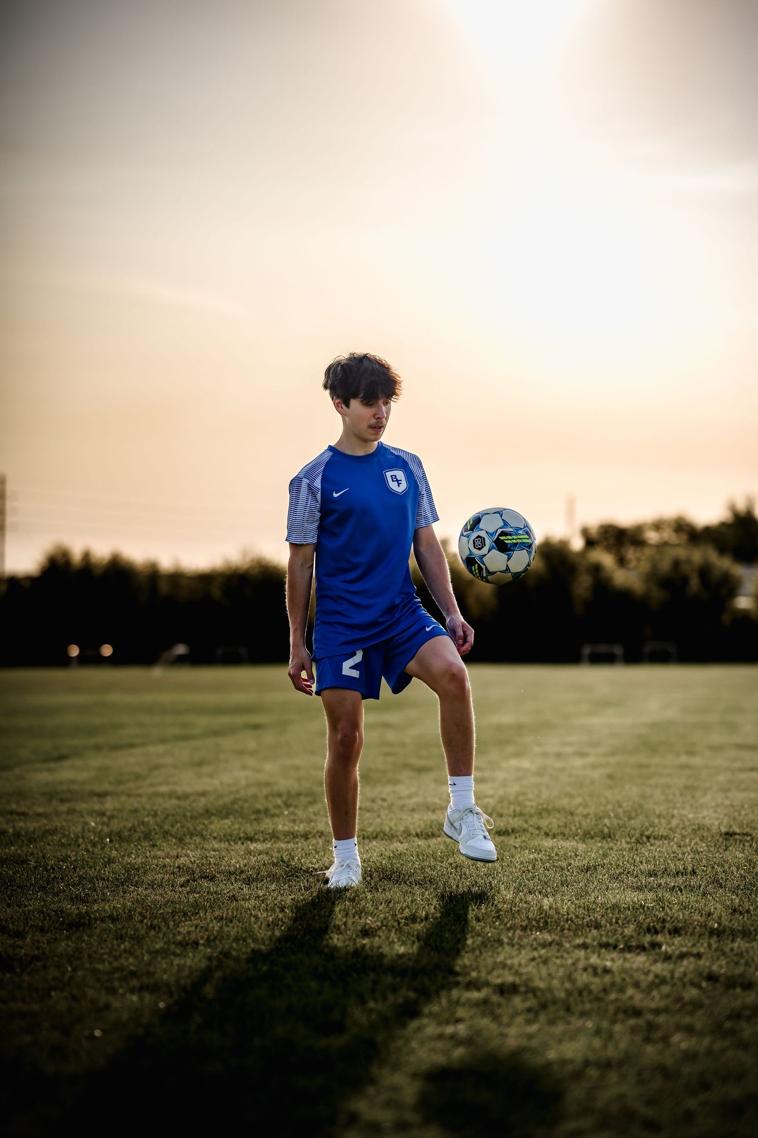 Teen soccer player in blue Nike uniform juggling ball on green field at sunset.
