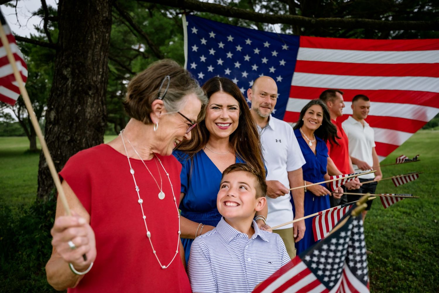Multi-generational family holding American flags outdoors with large US flag backdrop celebrating patriotic holiday.