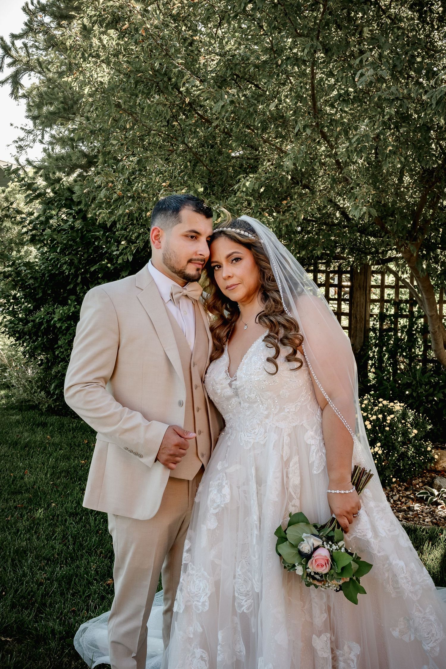 Bride in lace gown with veil and groom in tan suit pose together in lush garden setting.