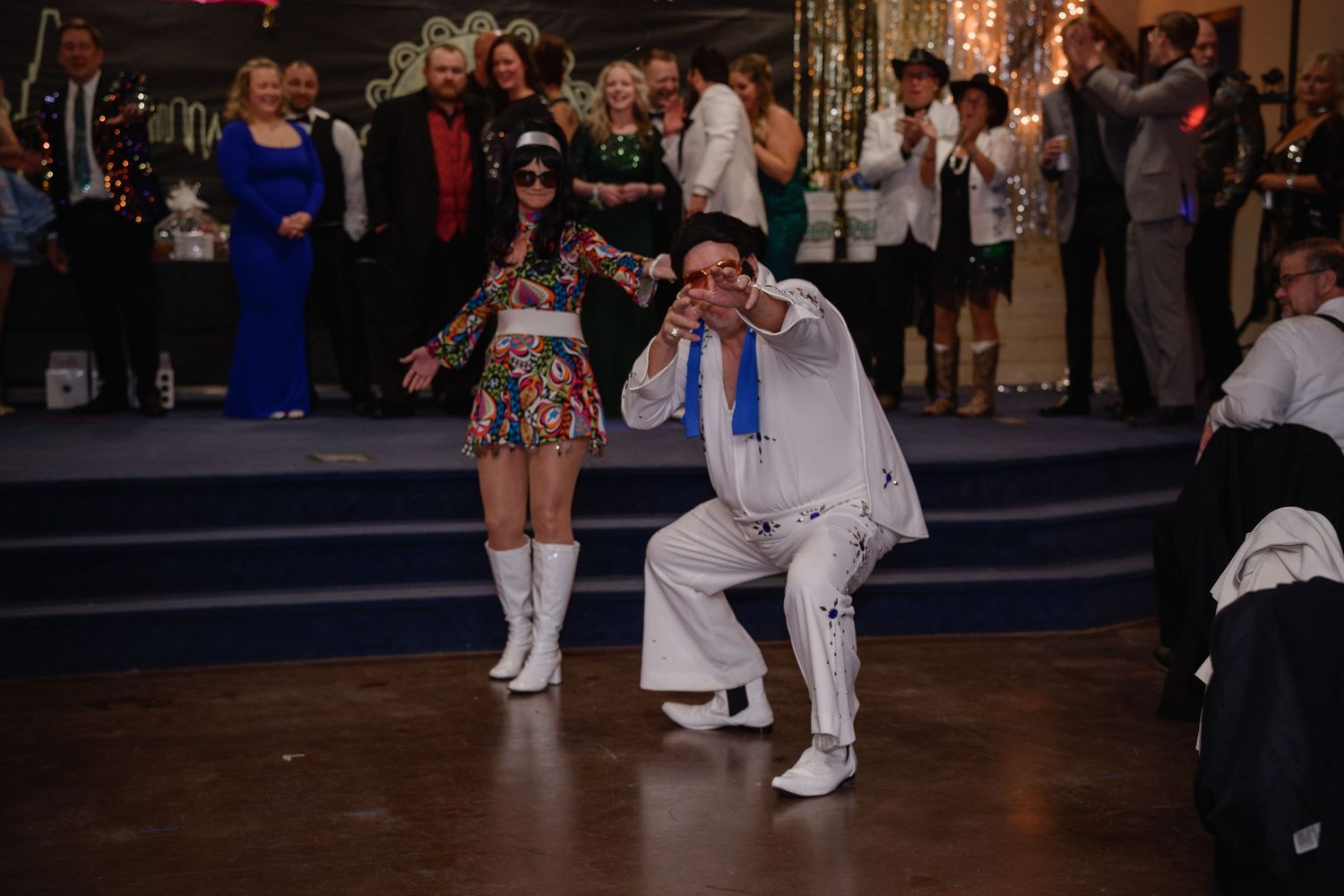 Elvis impersonator performing on dance floor with woman in colorful outfit at lively party event.