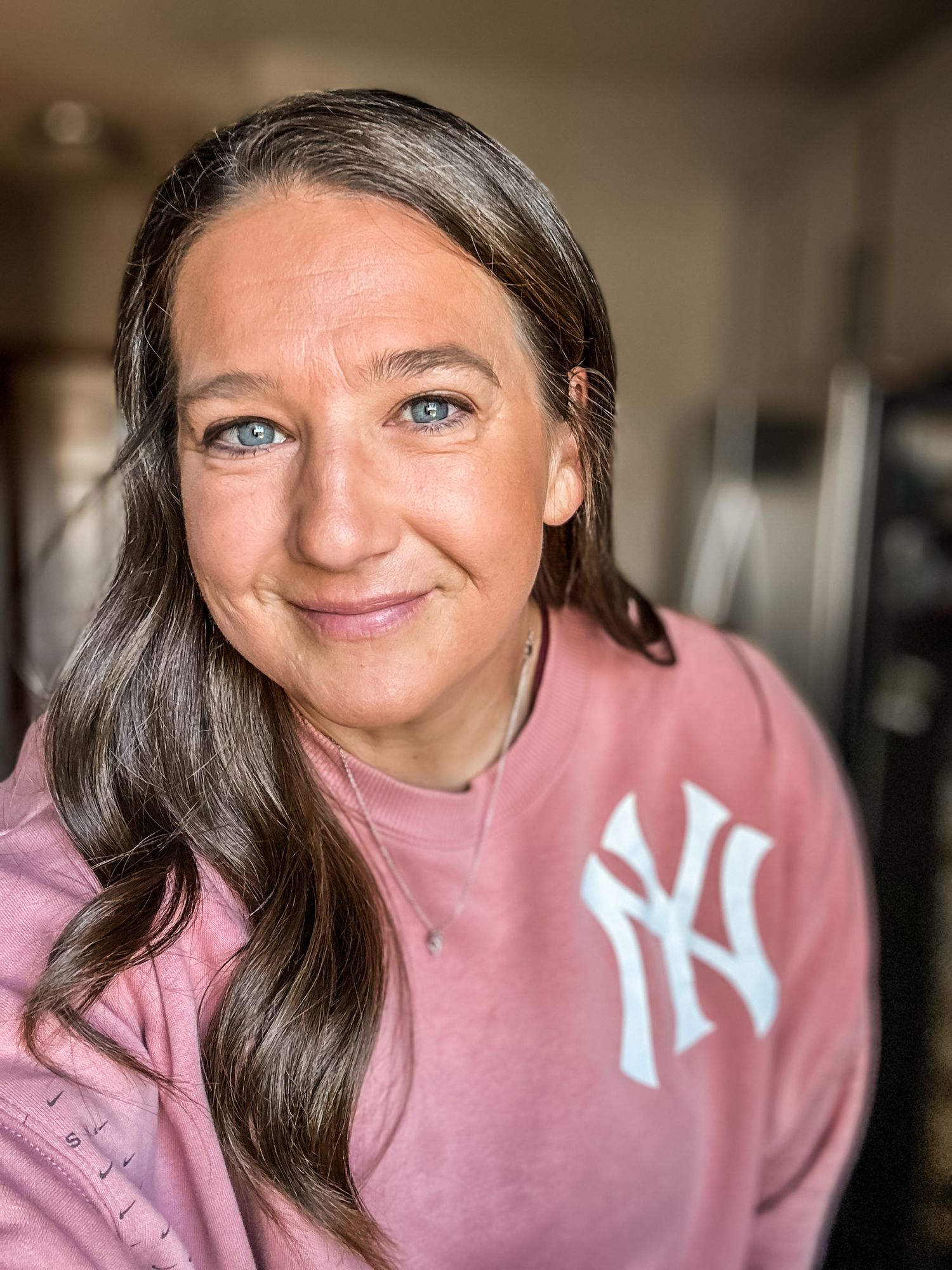 Smiling woman with blue eyes wearing pink New York Yankees sweatshirt in casual portrait.