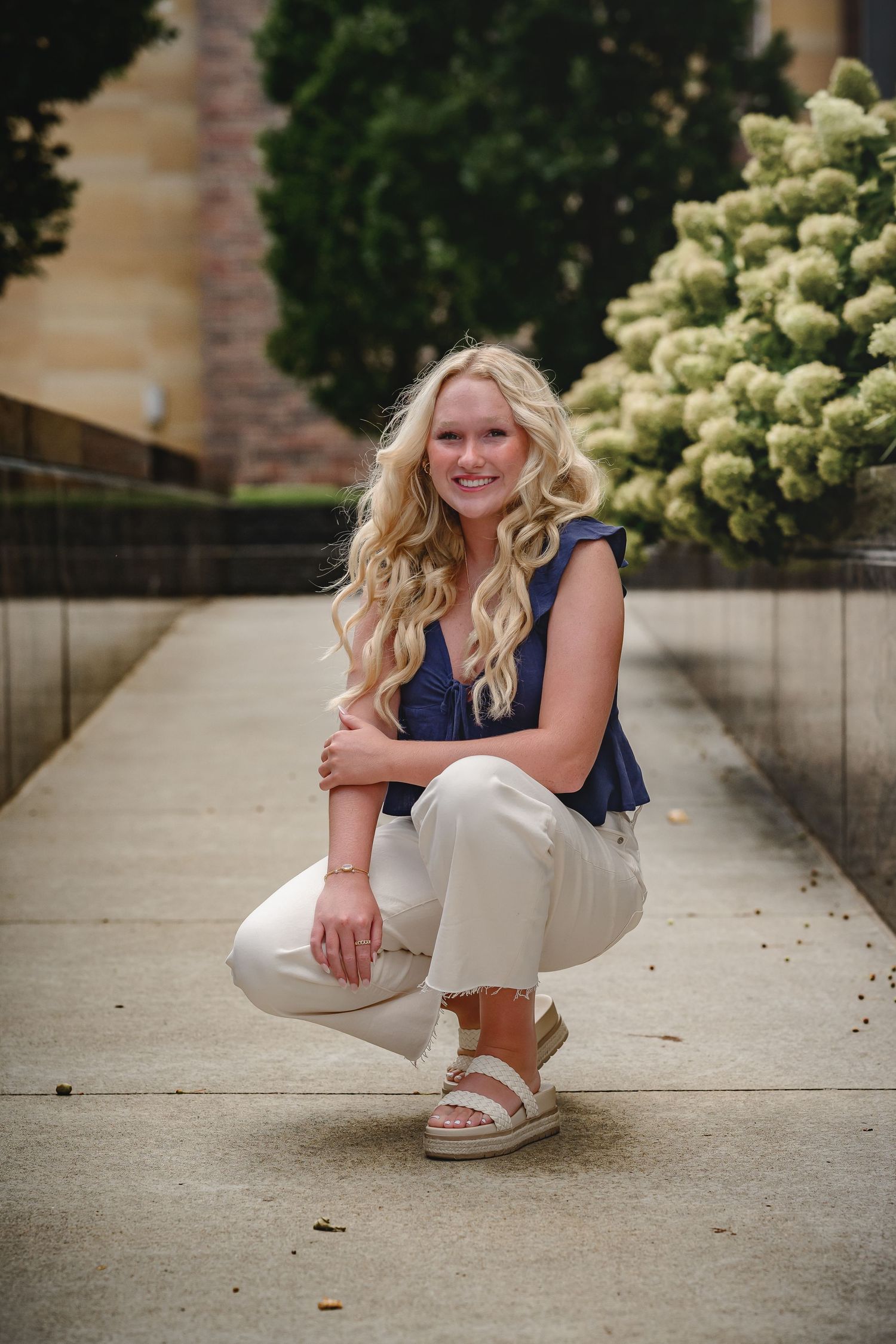 Smiling blonde woman crouching outdoors in navy top and white pants near flowering bushes.