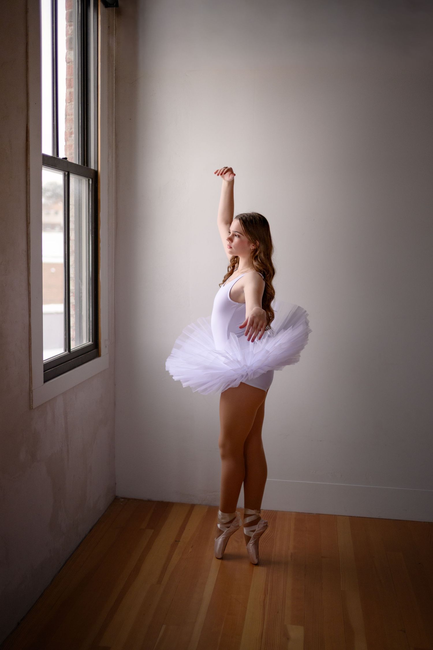 Ballerina in white tutu performing on pointe shoes in a bright studio near a large window.