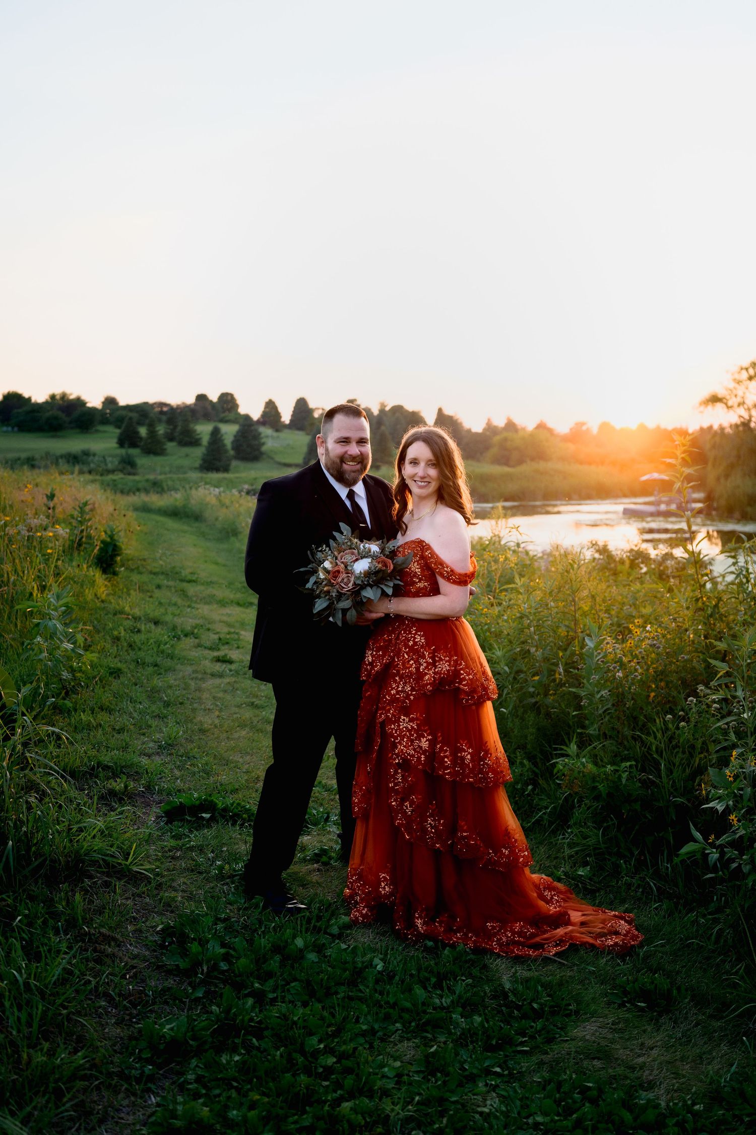 Couple poses at sunset, bride in red floral gown holding bouquet, groom in black suit, lush green landscape backdrop.
