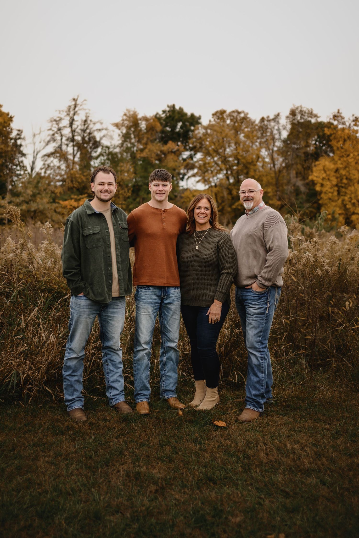 Family of four smiling together in autumn field surrounded by colorful fall foliage and tall grasses.