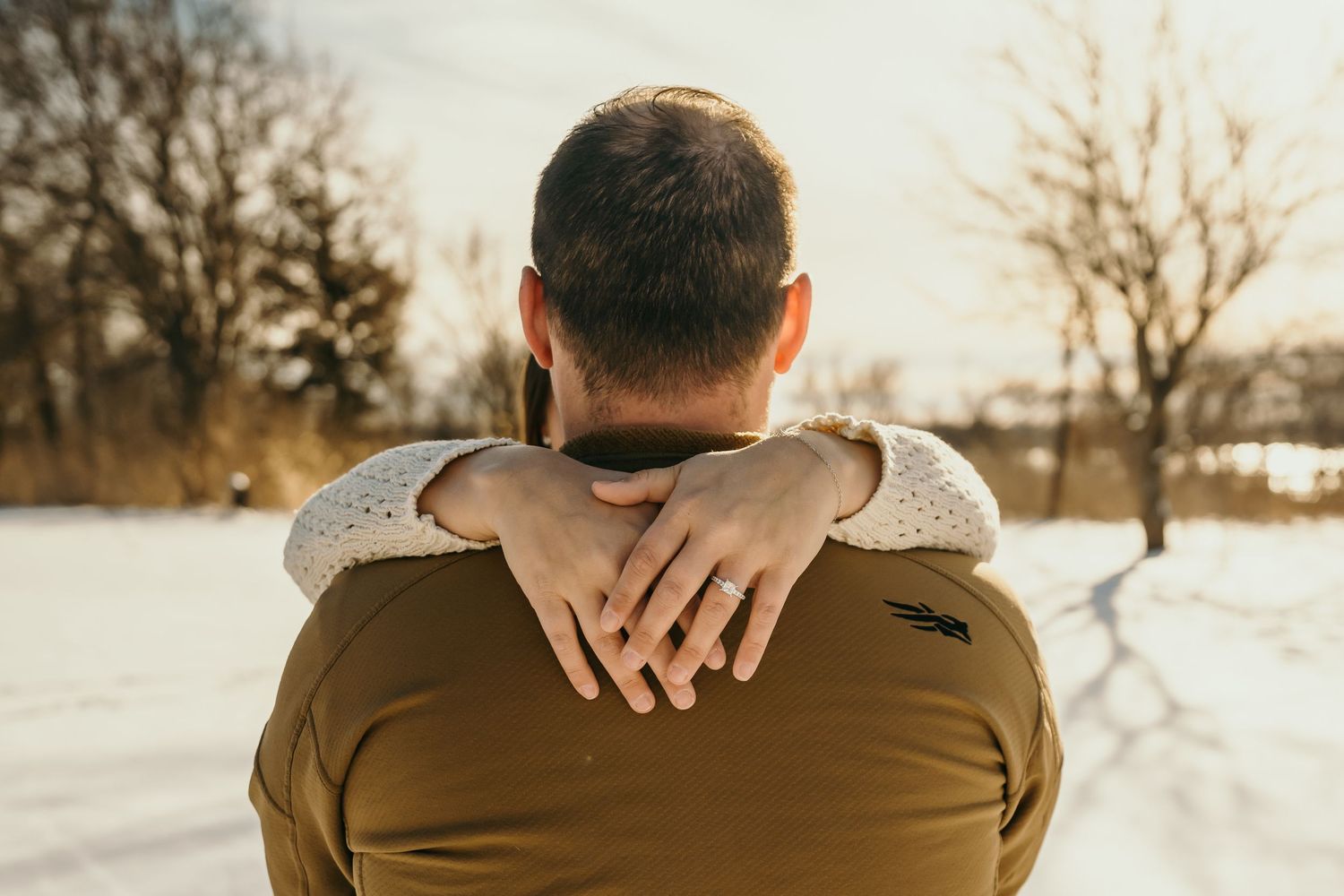 Romantic couple embrace from behind in snowy winter landscape, woman wearing engagement ring.