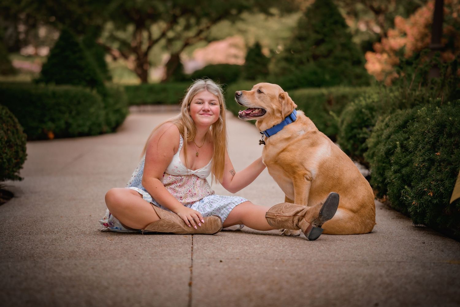 Smiling blonde woman sitting on pavement with golden Labrador Retriever in garden setting.