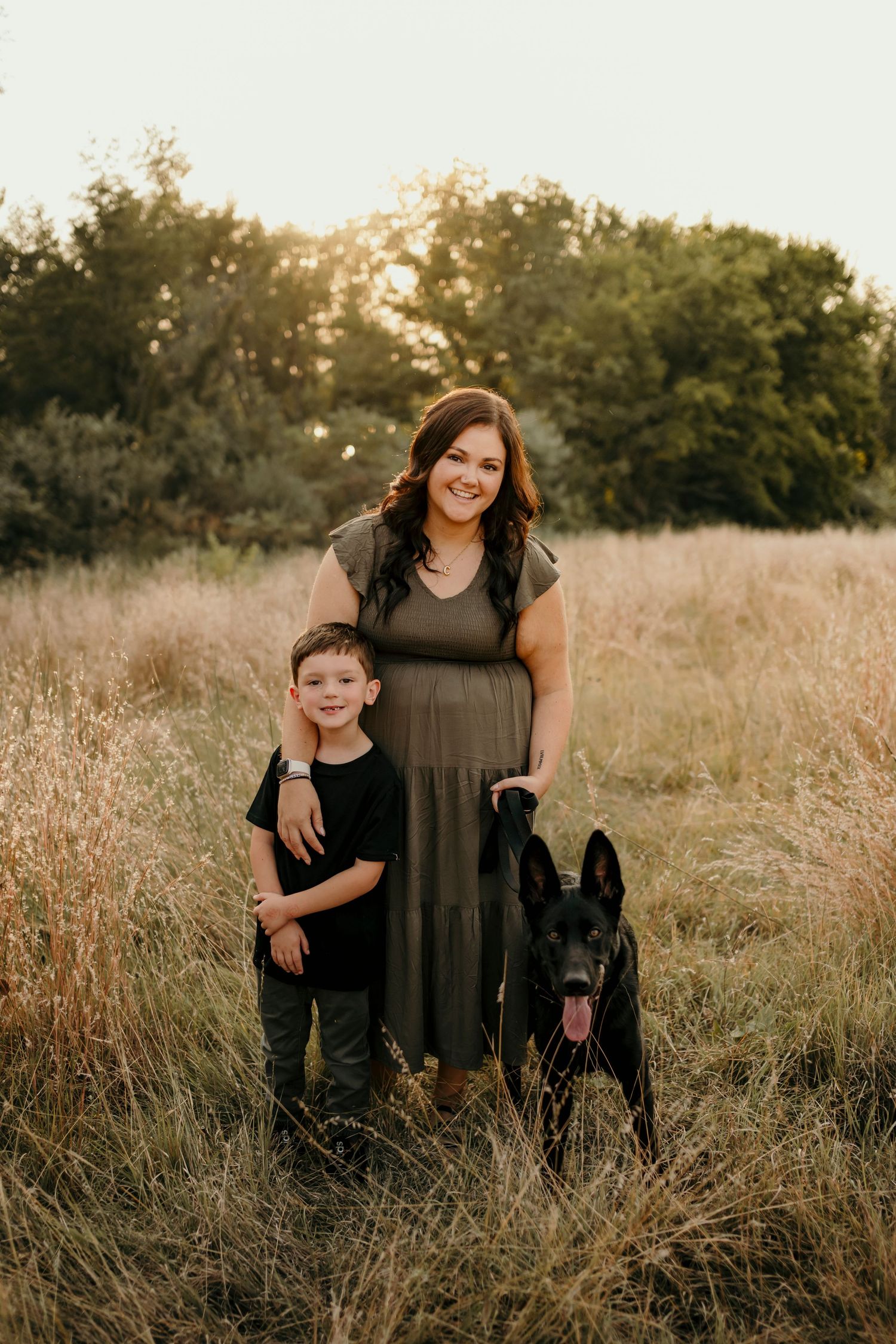 Smiling woman in olive dress standing with young boy and black German Shepherd in field.