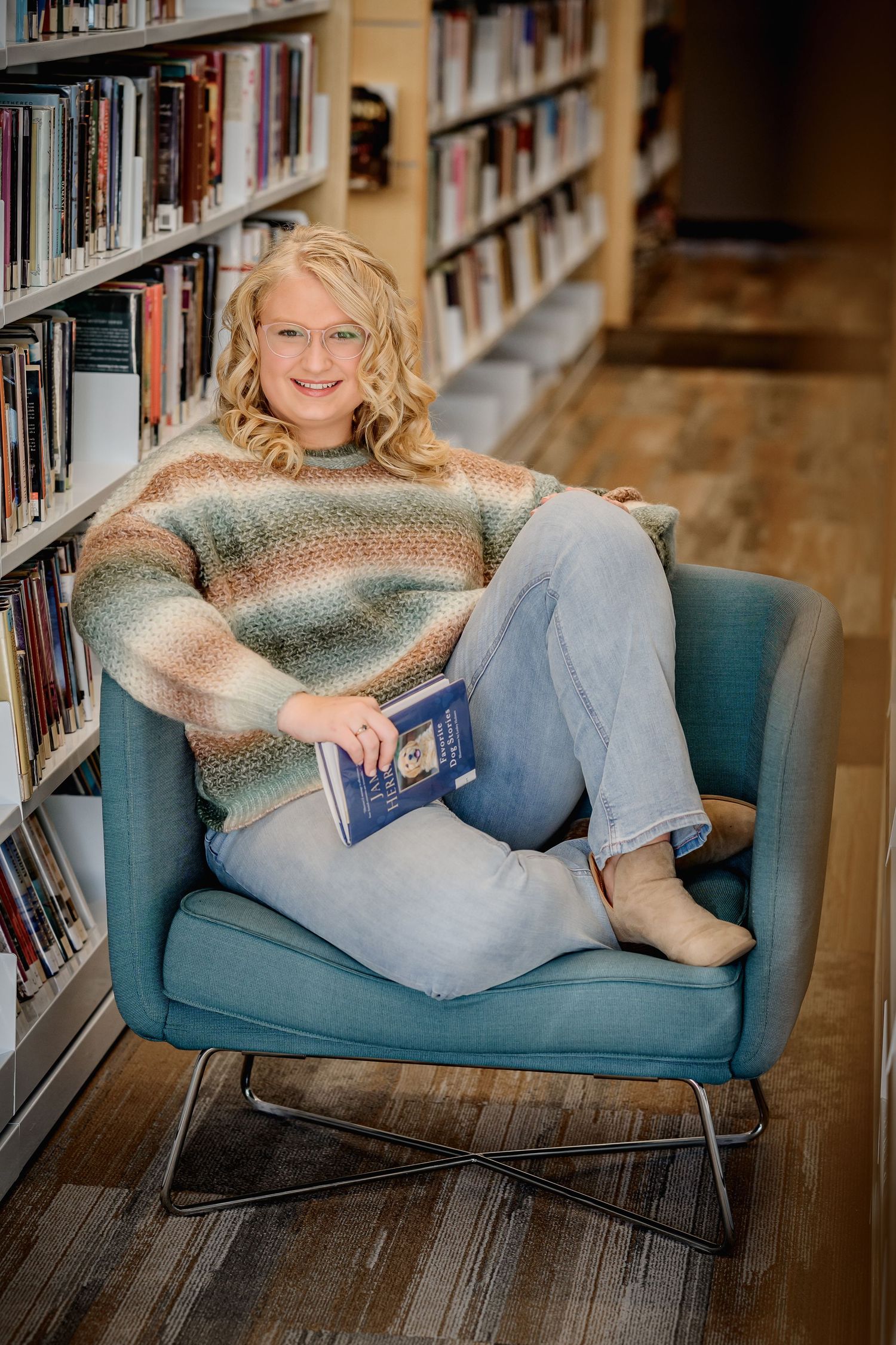 Smiling blonde teen girl sits in teal chair in library holding a book surrounded by bookshelves.