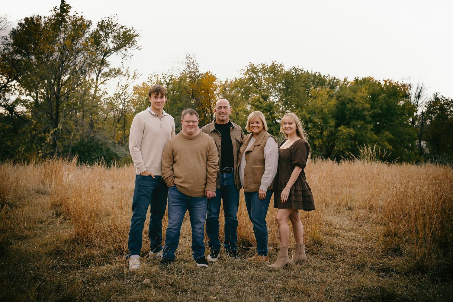 Family of five smiling together in an autumn field with golden grass and colorful fall foliage.