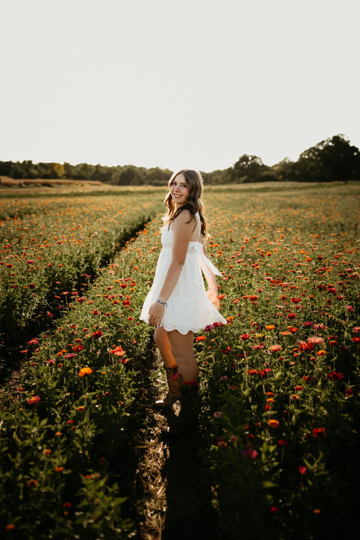 A smiling young woman in a white dress walks through rows of colorful blooming flowers at sunset.