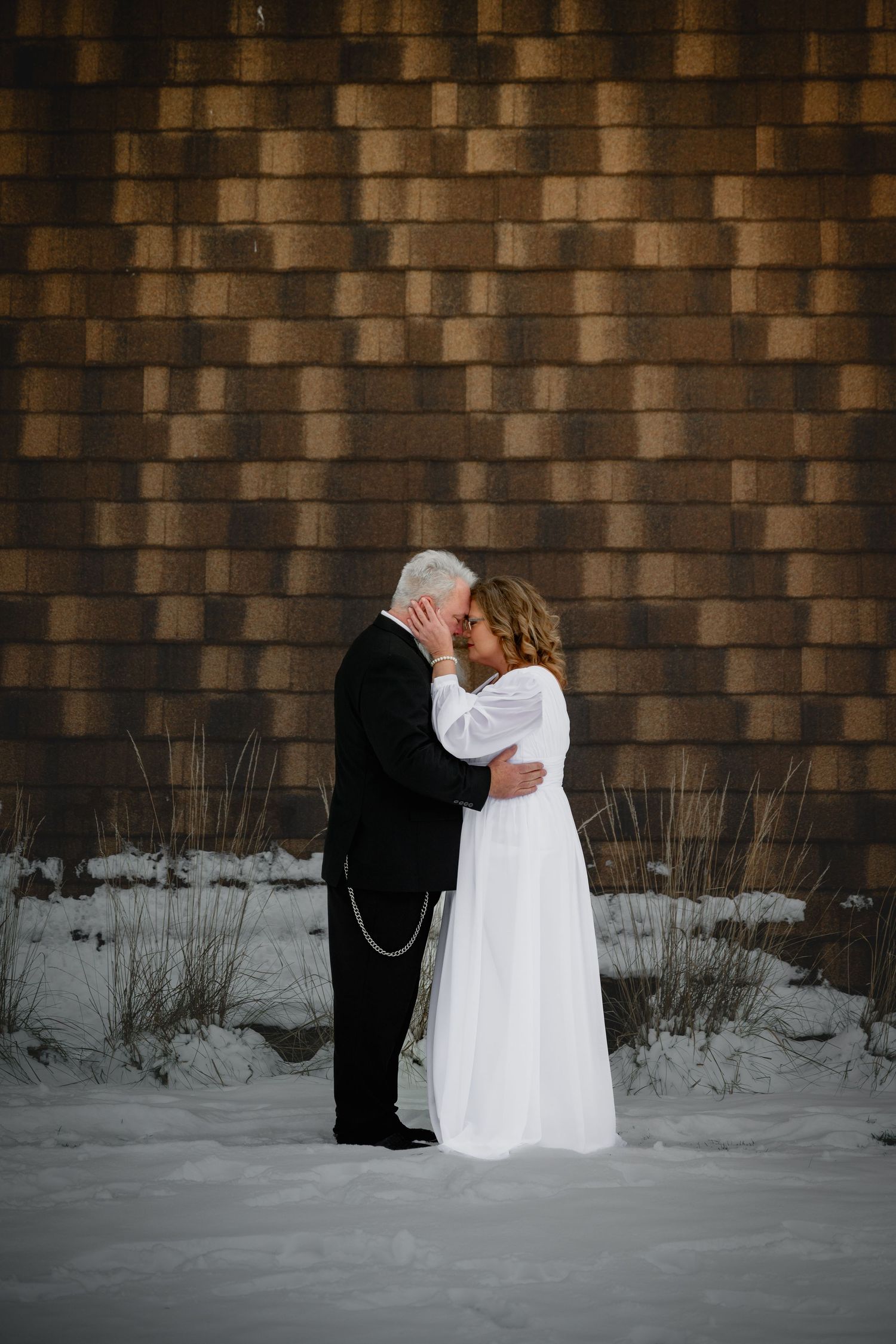 Bride and groom sharing a romantic kiss in the snow against a rustic brick wall in winter.