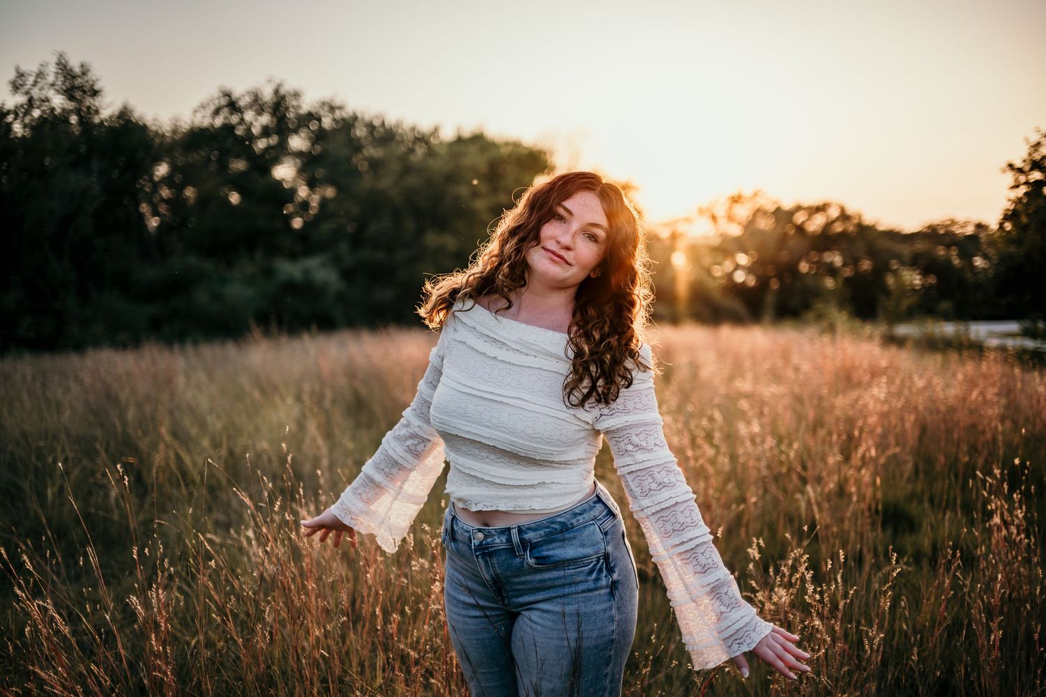 Young woman with curly hair in white lace top and jeans standing in golden field at sunset.