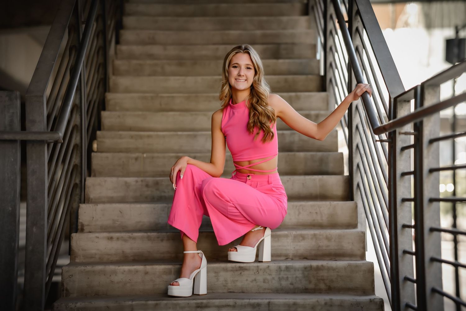 Smiling young woman in pink outfit posing on concrete staircase with metal railing.
